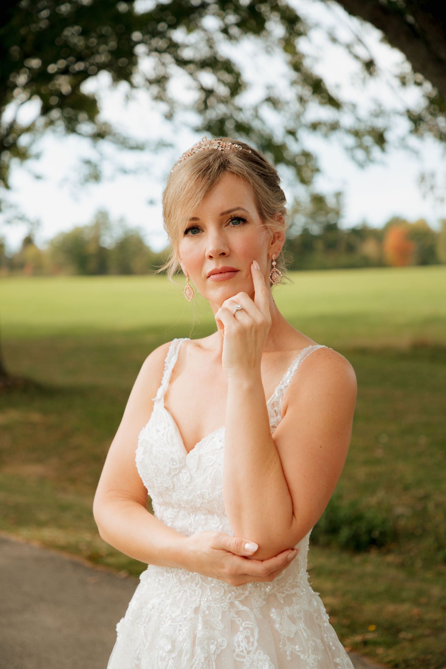 Elegant bride in a lace gown and tiara poses thoughtfully with hand near chin in an outdoor park setting.