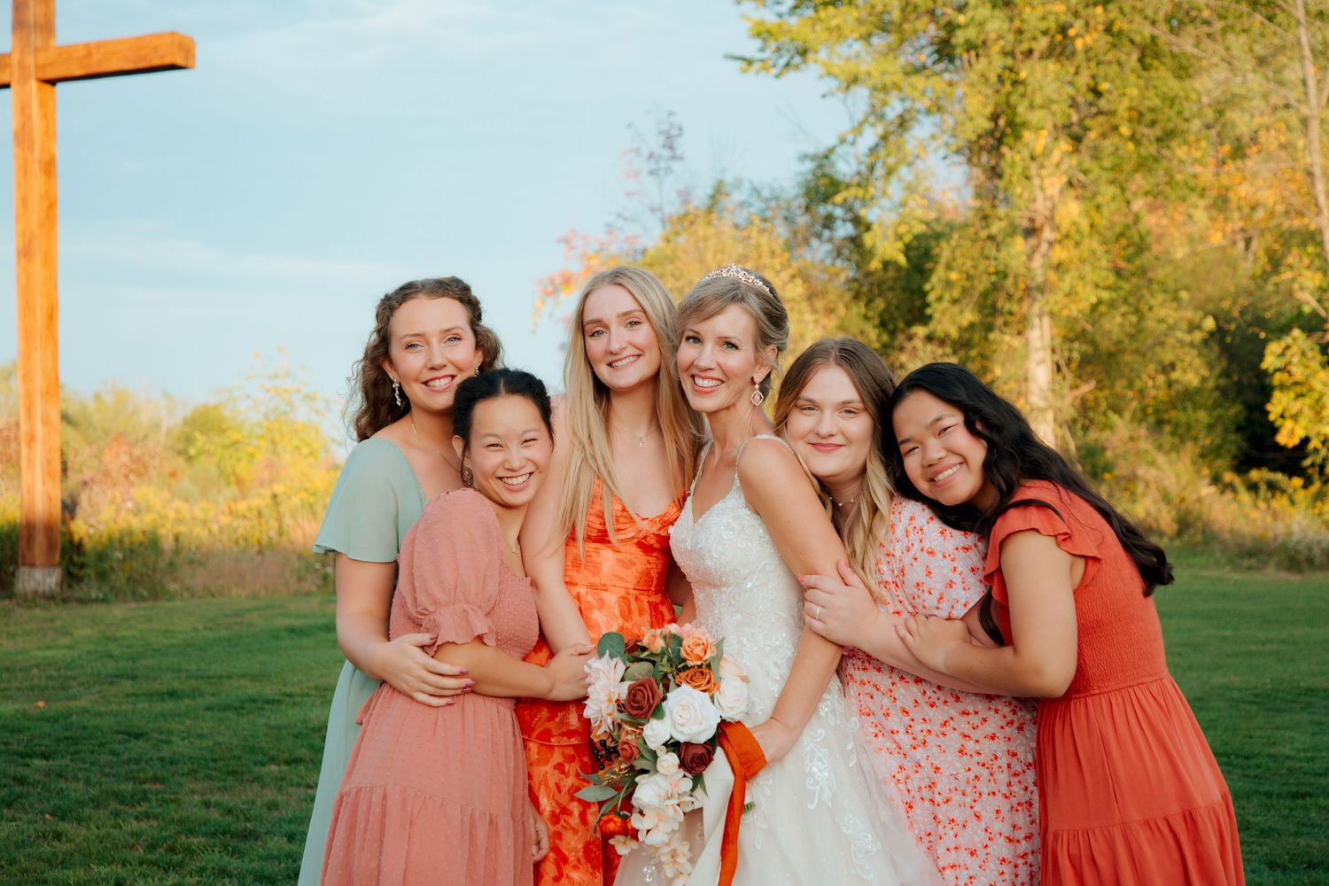Bride in white gown smiling with four bridesmaids in colorful dresses at an outdoor wedding.
