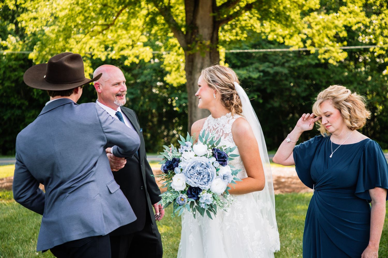 Bride smiling with white floral bouquet greets guests alongside groom in cowboy hat at sunny outdoor wedding reception.