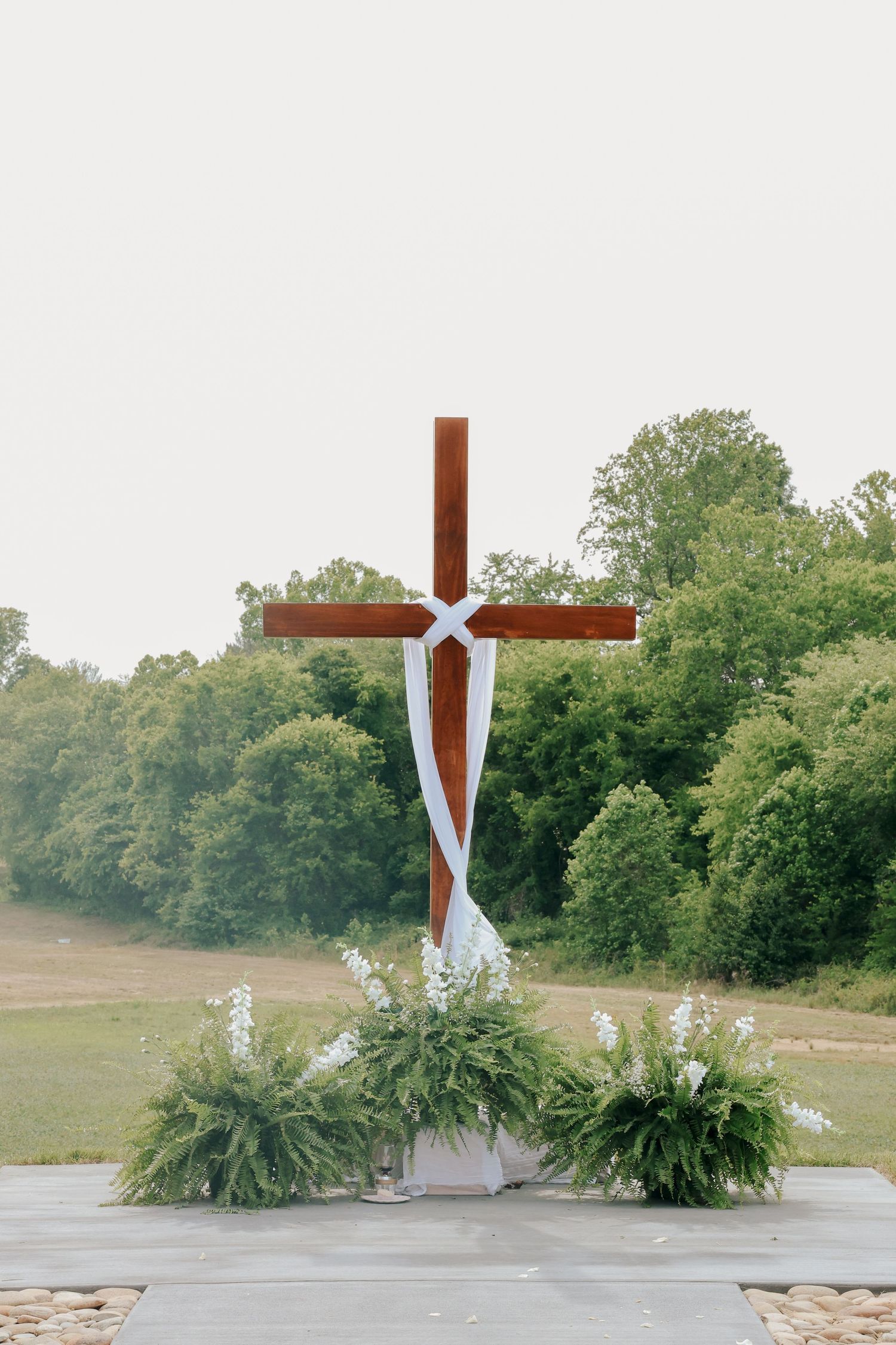 Wooden cross decorated with white ribbon and greenery floral arrangements at an outdoor wedding ceremony.