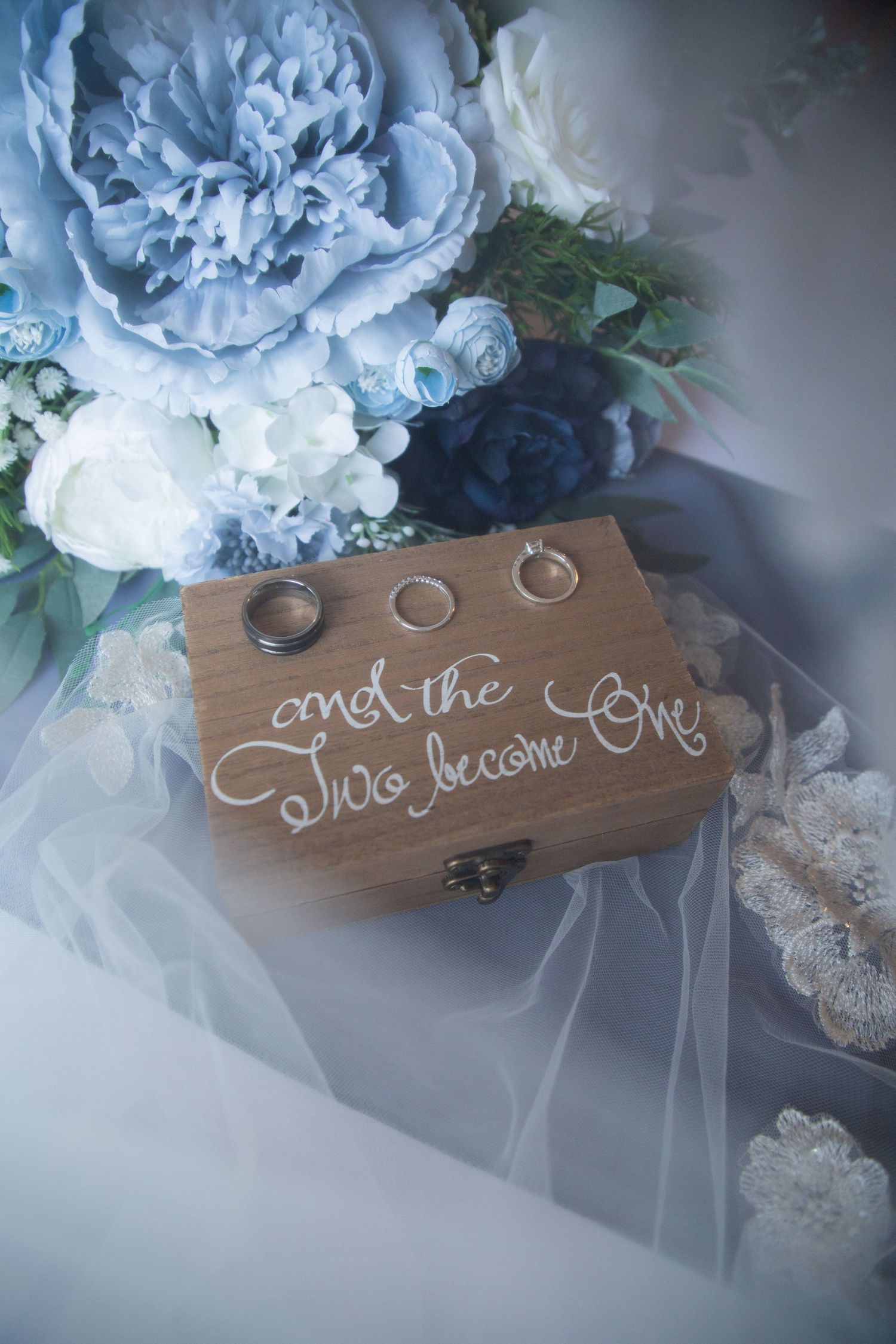 Wooden ring box inscribed with 'and the two become one' surrounded by blue and white wedding flowers.