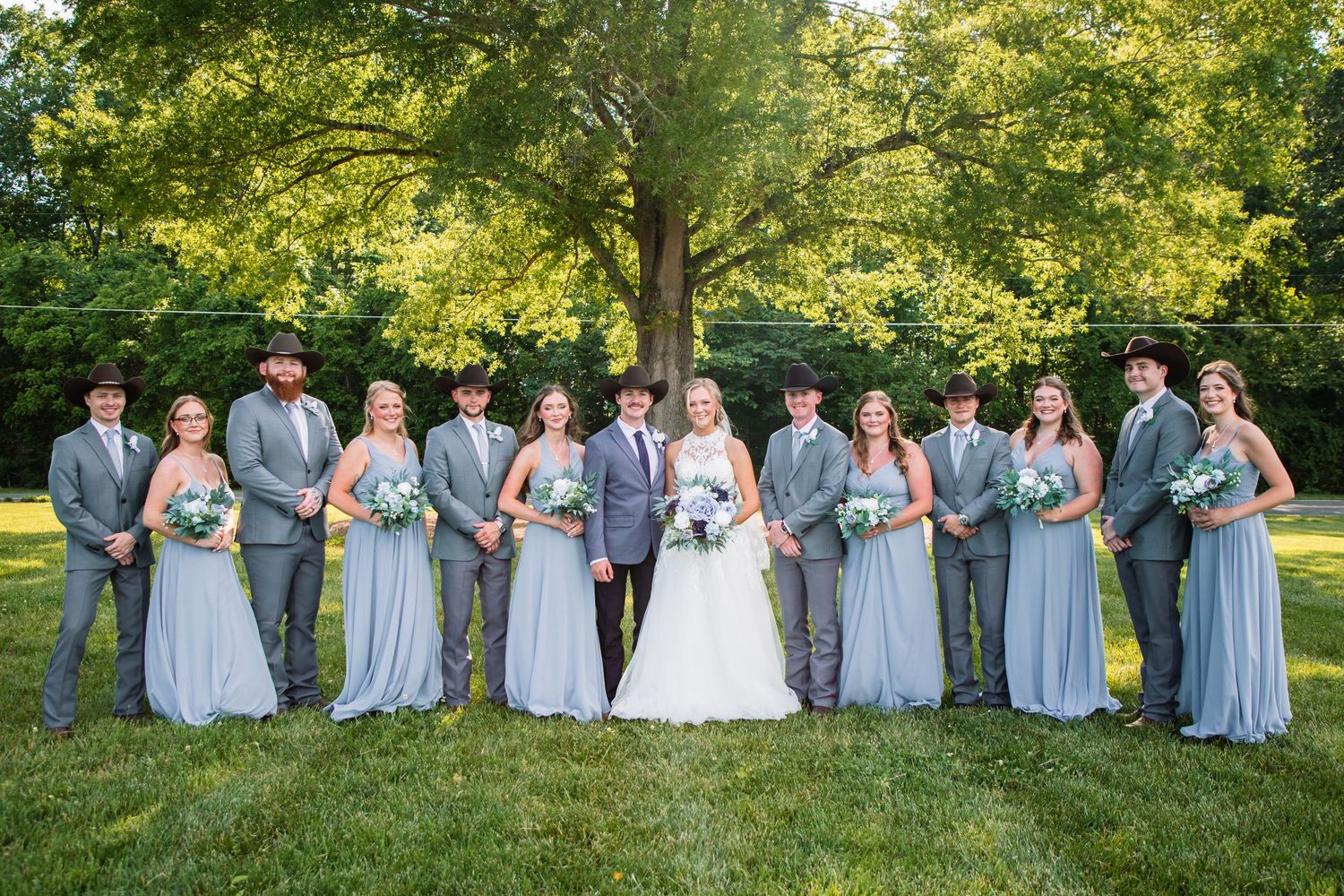 Wedding party smiles together outdoors, bridesmaids in dusty blue gowns and groomsmen in gray suits with cowboy hats.