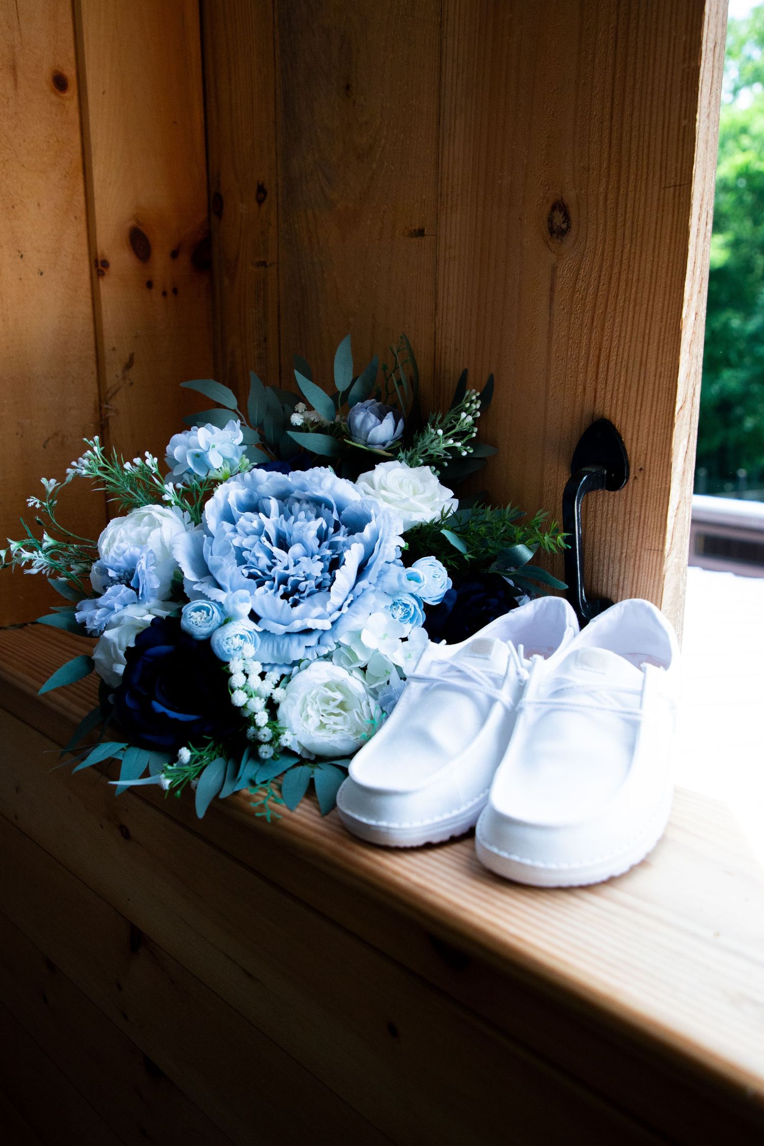 Blue and white wedding bouquet with white groom's shoes on wooden shelf in rustic setting.