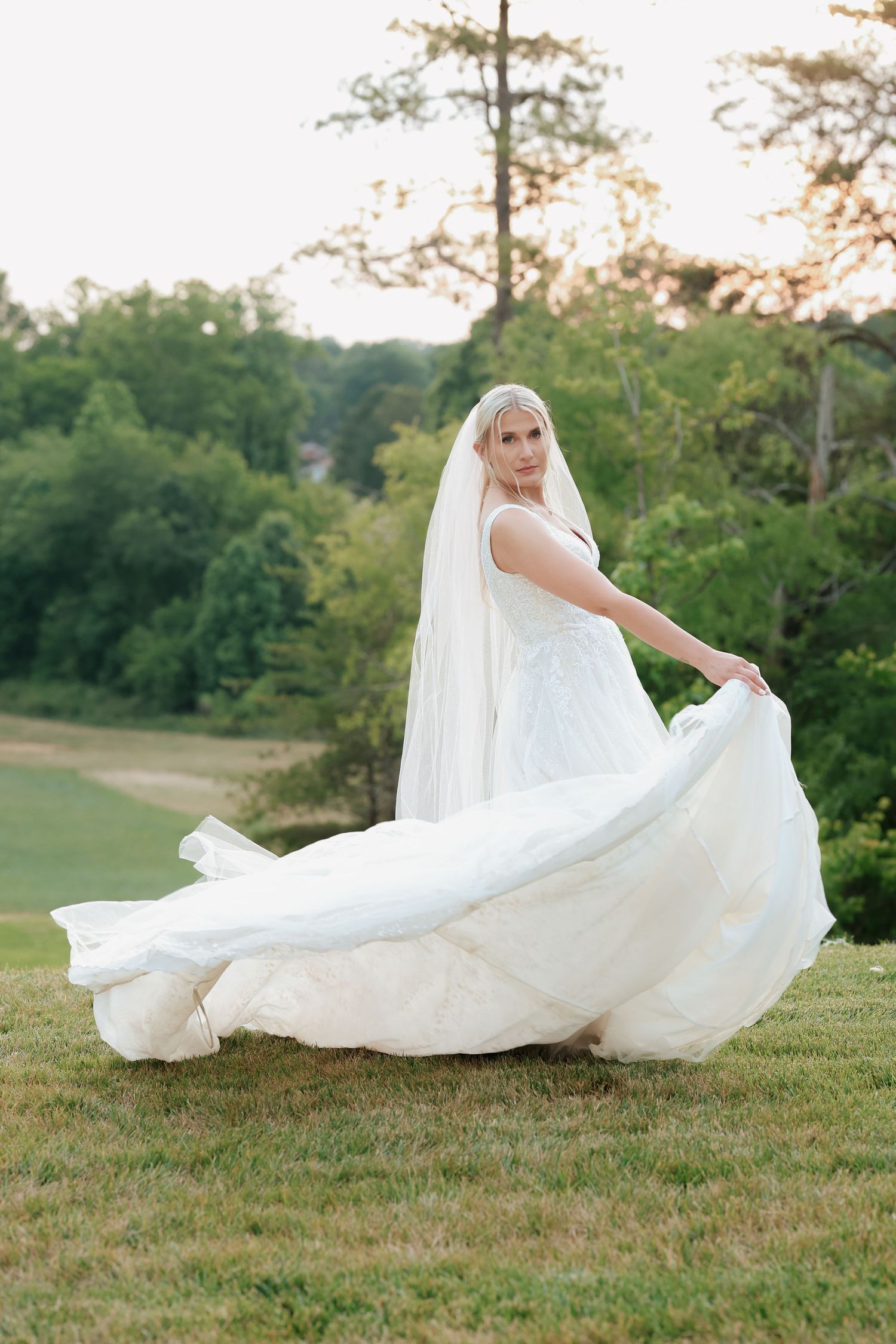 Bride in flowing white gown and veil twirling on green lawn at outdoor wedding venue.