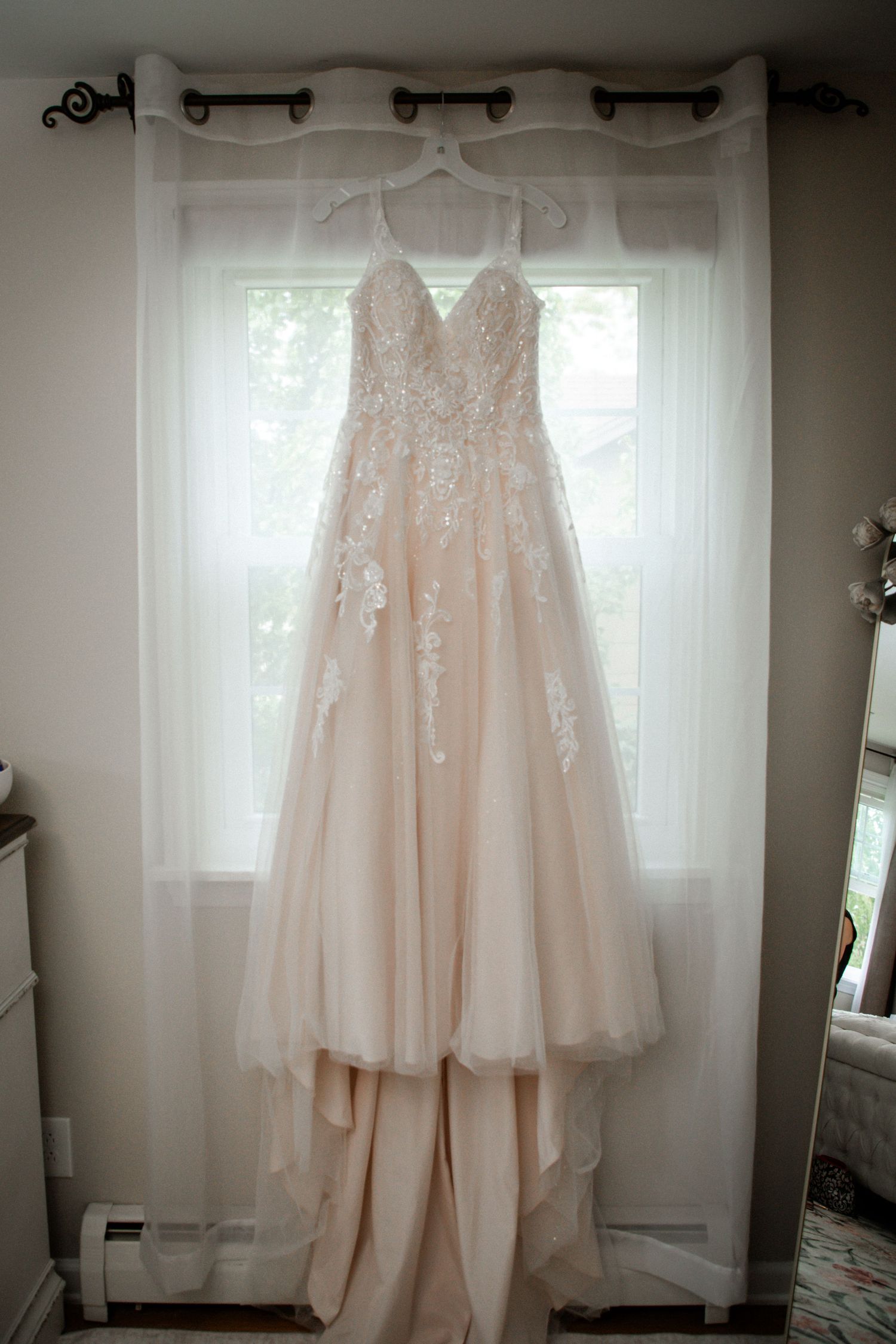 Blush lace wedding gown with floral appliques hanging by bright window before ceremony.