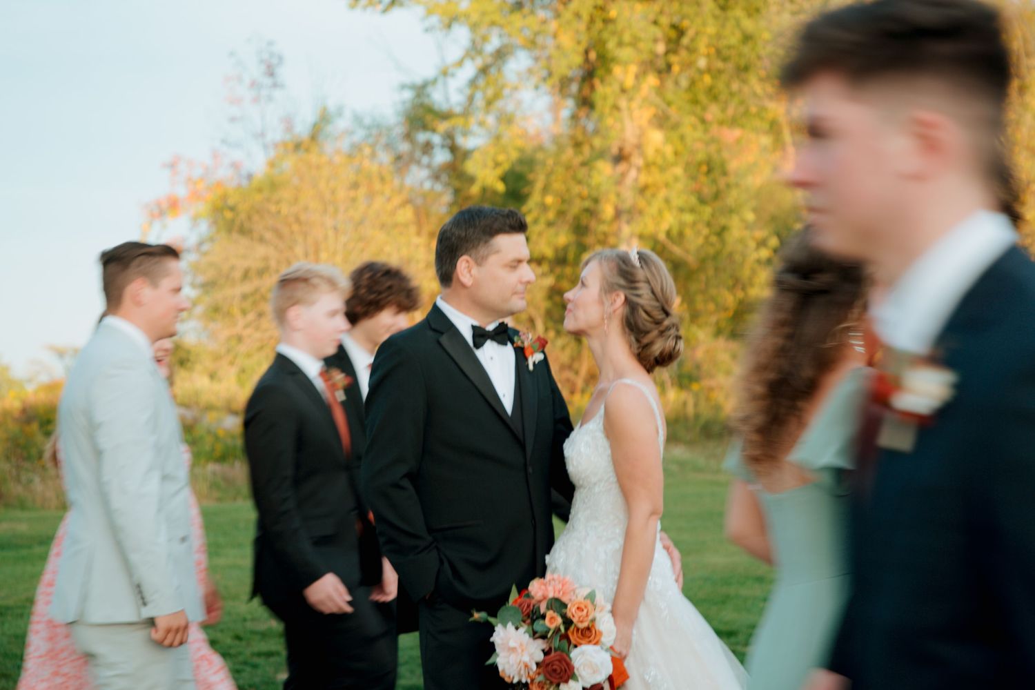 Groom in black tuxedo gazing at bride holding bouquet during outdoor fall wedding ceremony.