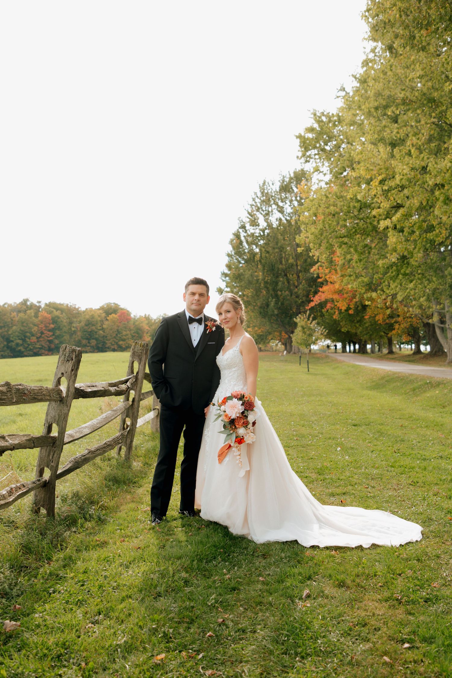 Bride and groom posing by a rustic wooden fence on a lush green hillside during an autumn wedding.
