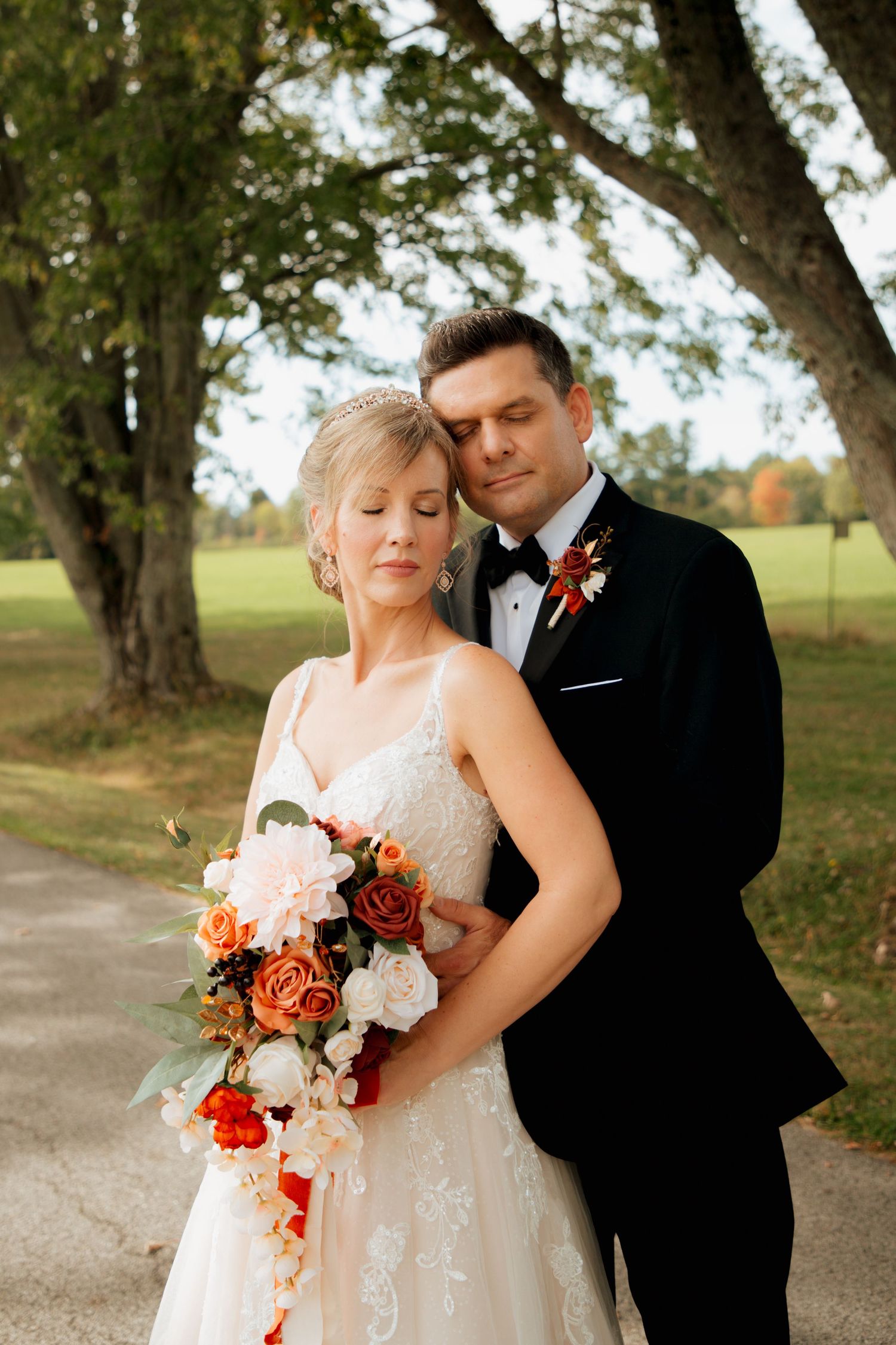 Groom tenderly embraces bride from behind as both close their eyes on a scenic tree-lined pathway.
