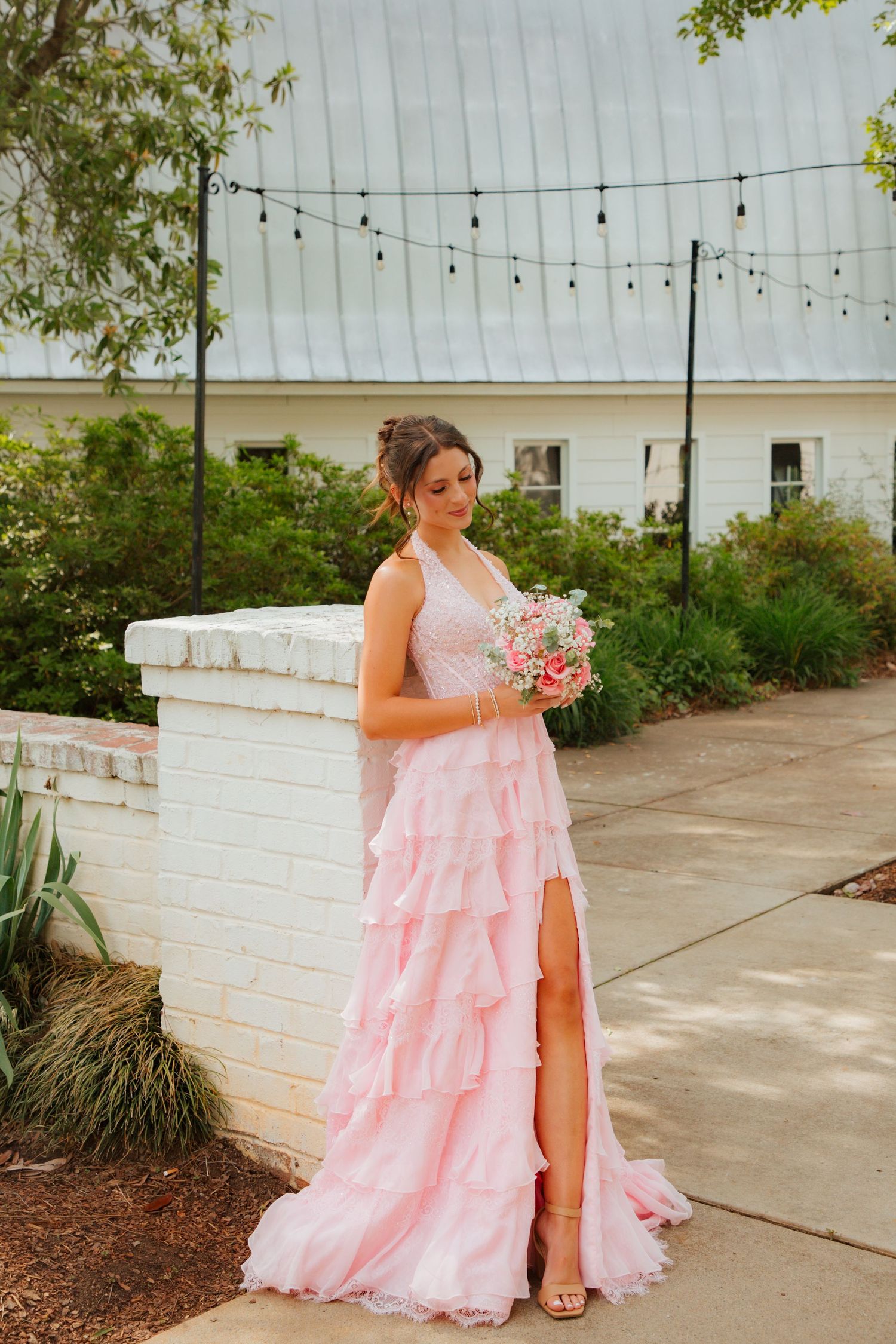 Elegant bride in layered pink ruffle gown holding bouquet, posing beside white brick wall with string lights above.