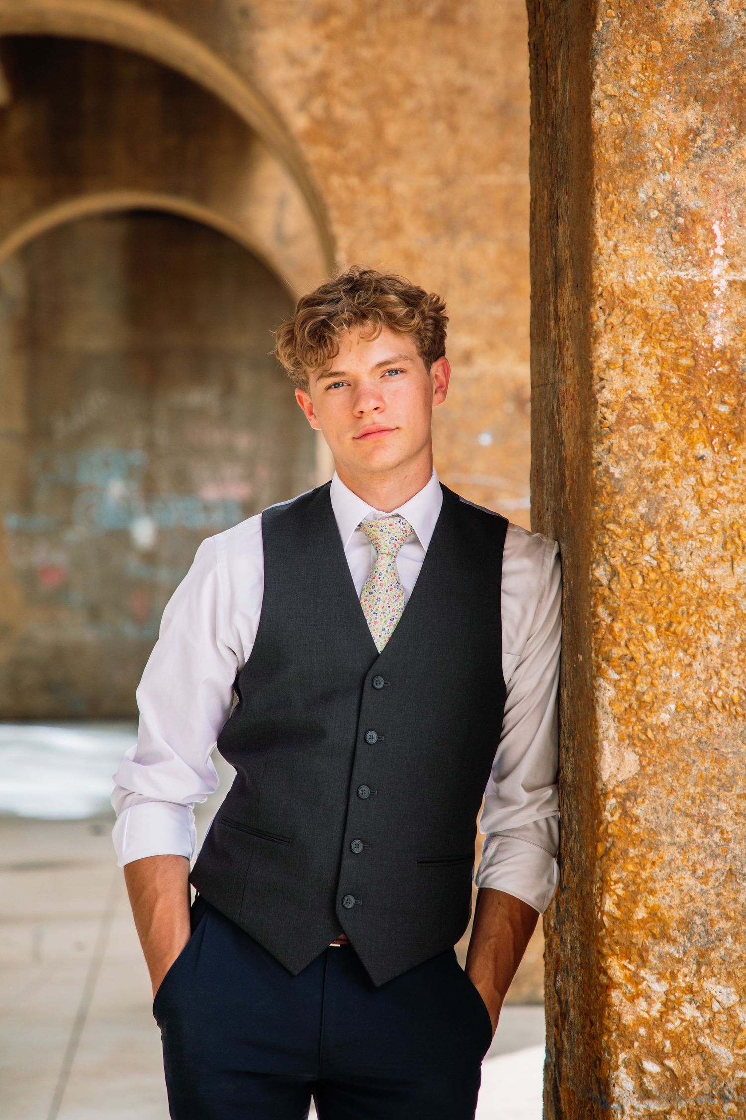 Stylish young man in black vest and gold tie leans against rustic stone column.