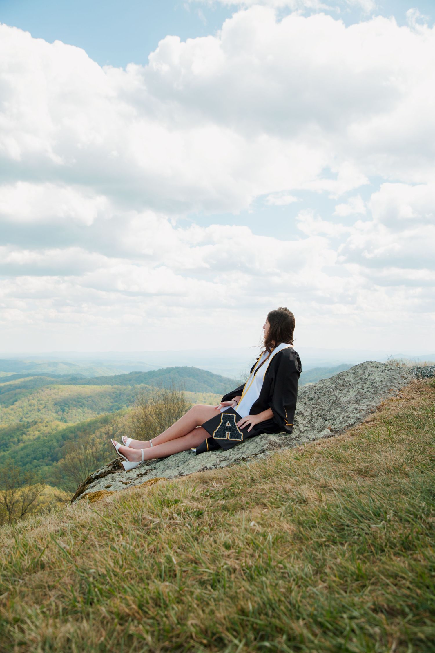 Graduate in black cap and gown sitting on rocky hillside overlooking stunning mountain landscape.