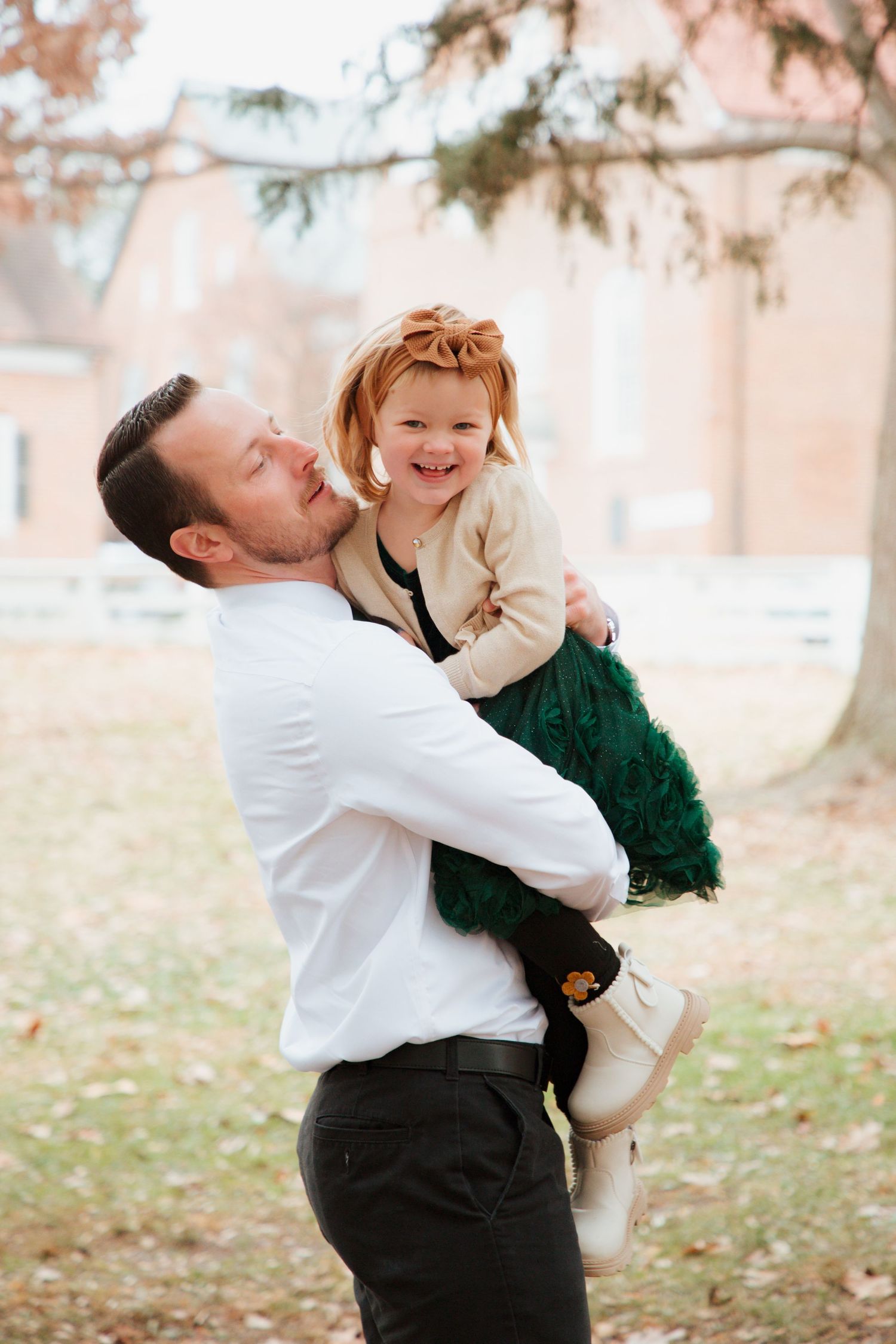 Father lifting laughing toddler girl in green dress outdoors during fall family photo session.