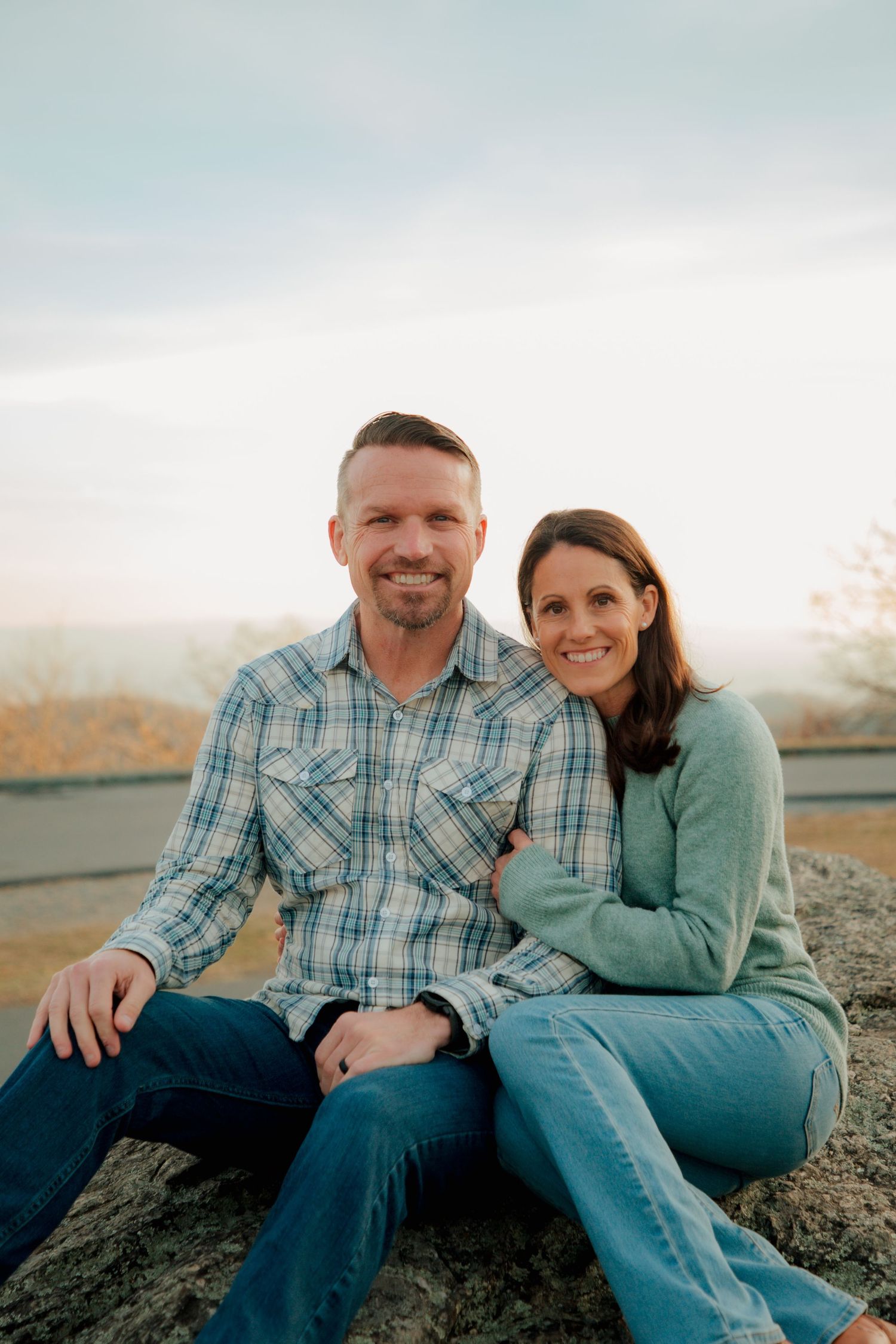 Cheerful couple sits closely on a rock with rolling hills and autumn landscape in background.