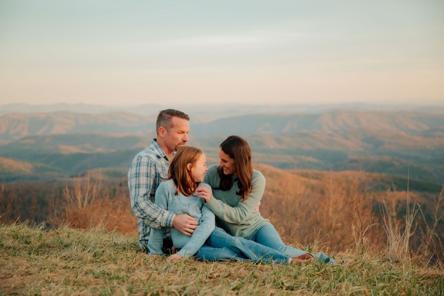 A loving family of three sits together on a grassy mountain overlook enjoying a warm sunset together.