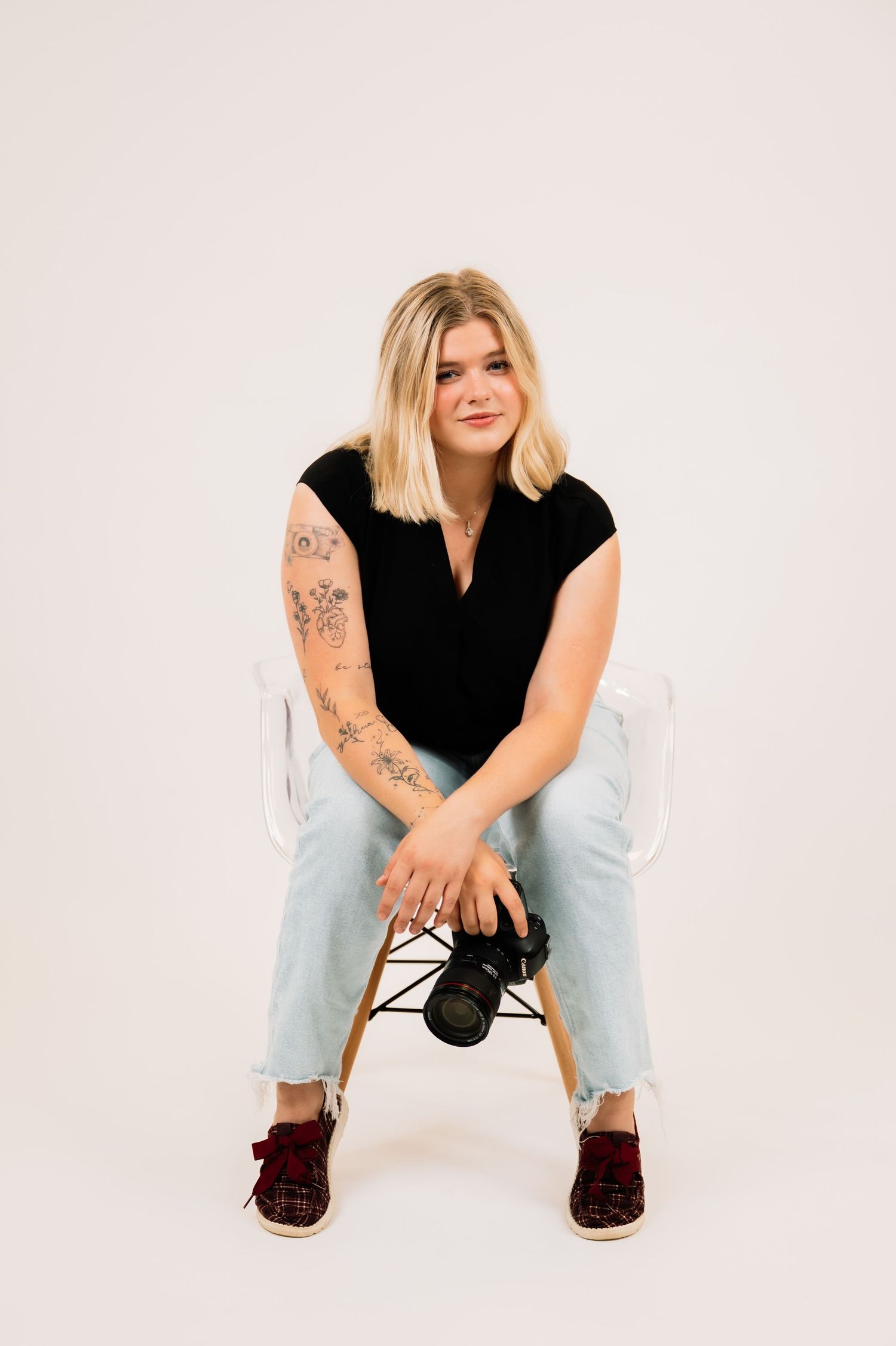 Tattooed blonde woman sitting in acrylic chair holding DSLR camera wearing black top and light jeans.