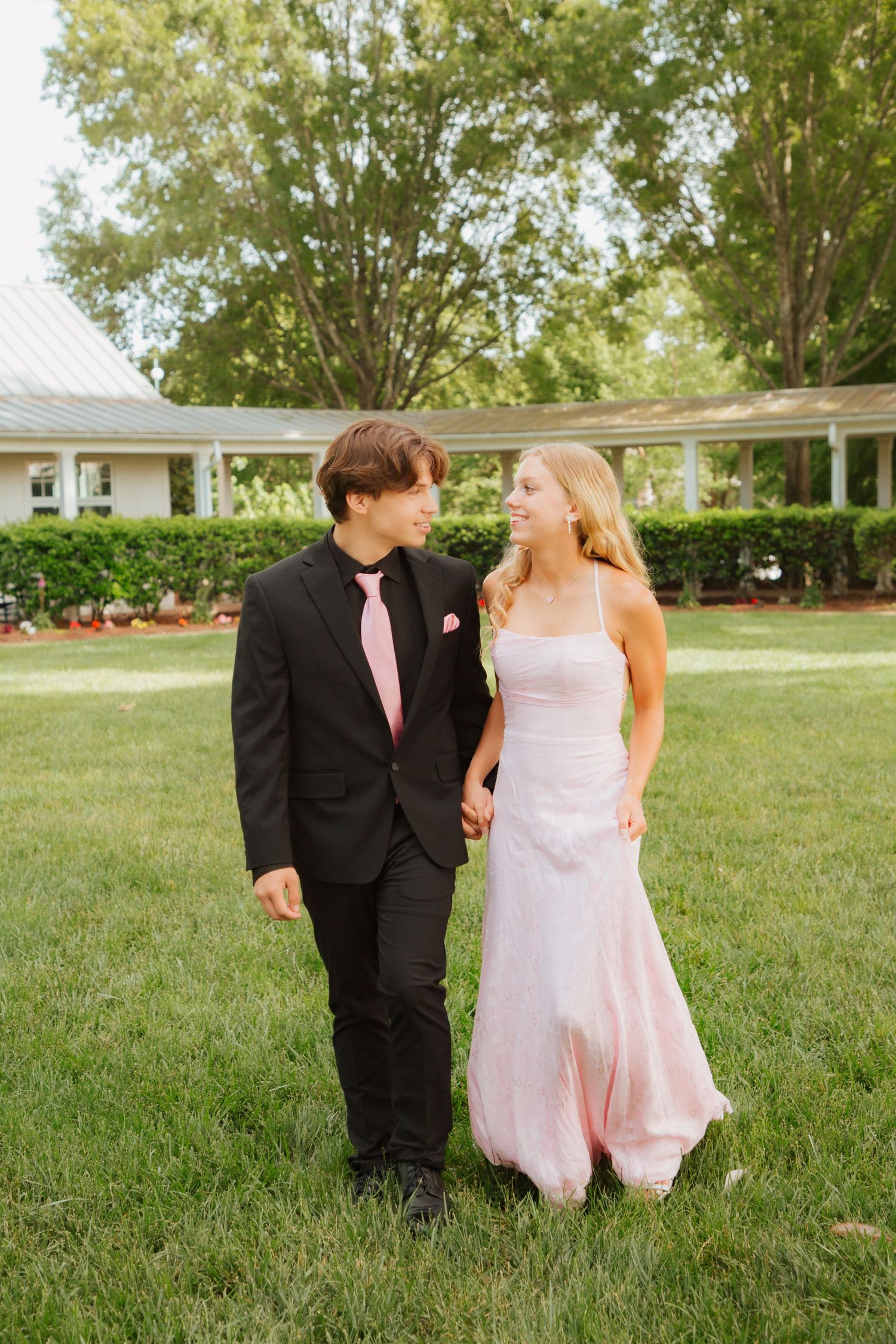 Teen prom couple holding hands and smiling at each other while walking on green lawn.