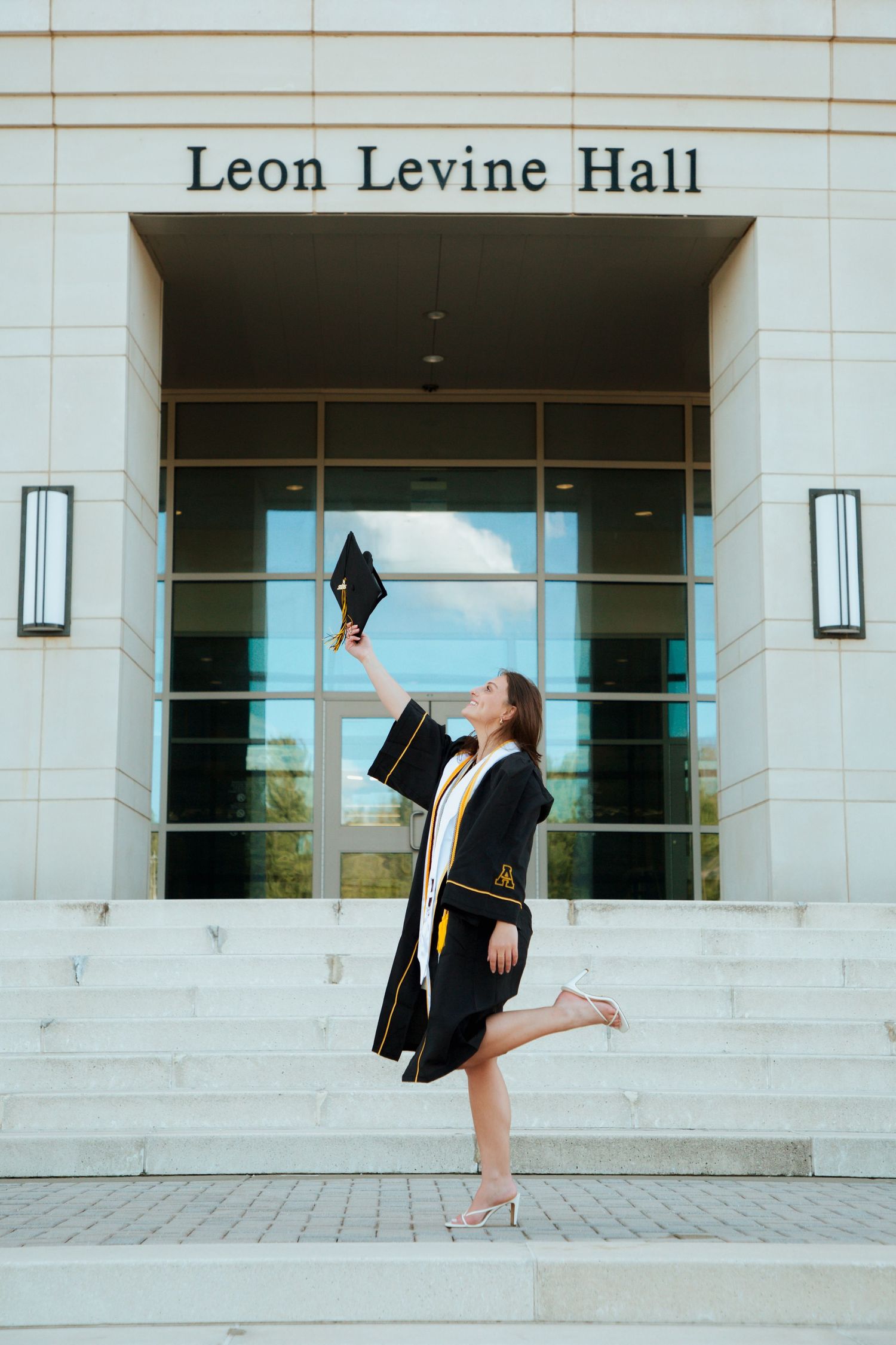 Joyful graduate in black gown raises cap outside Leon Levine Hall on one leg.