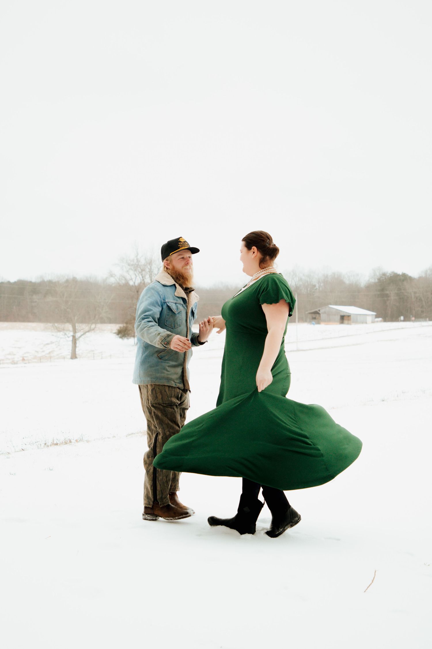 Couple dancing in snowy field, woman in green dress twirling, man in denim jacket and cap.