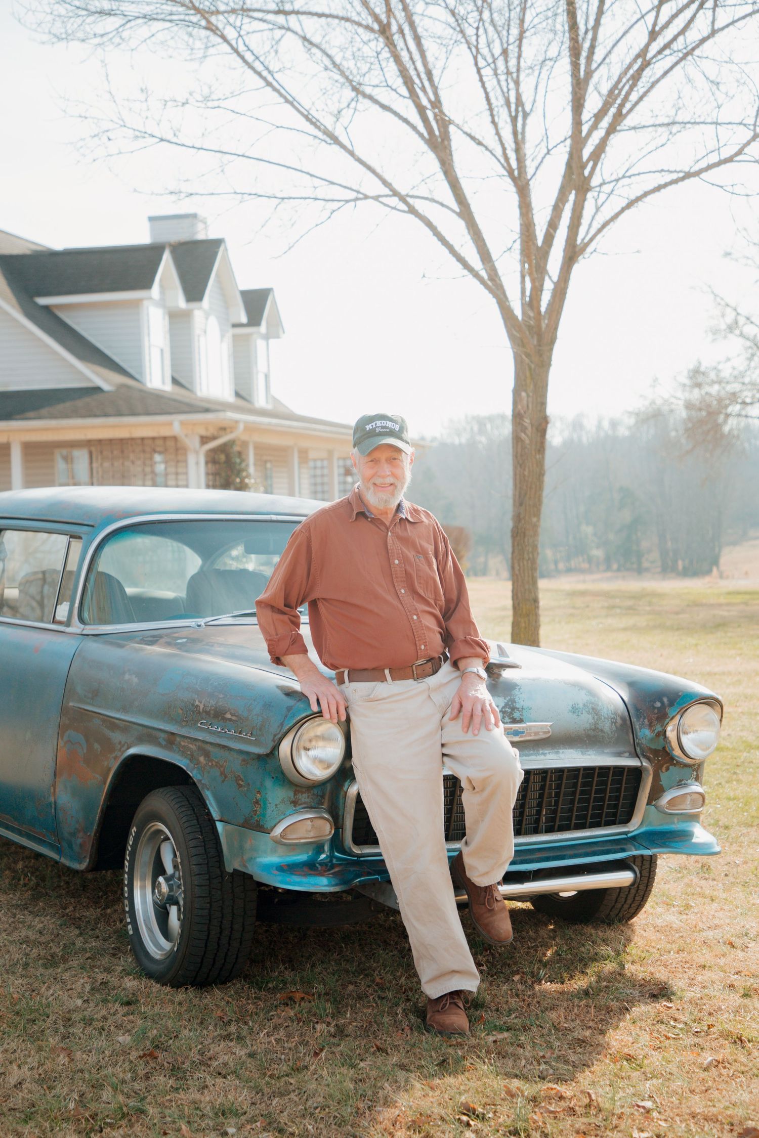 Senior man in a rust shirt and cap leaning proudly against a vintage teal 1955 Chevrolet classic car.