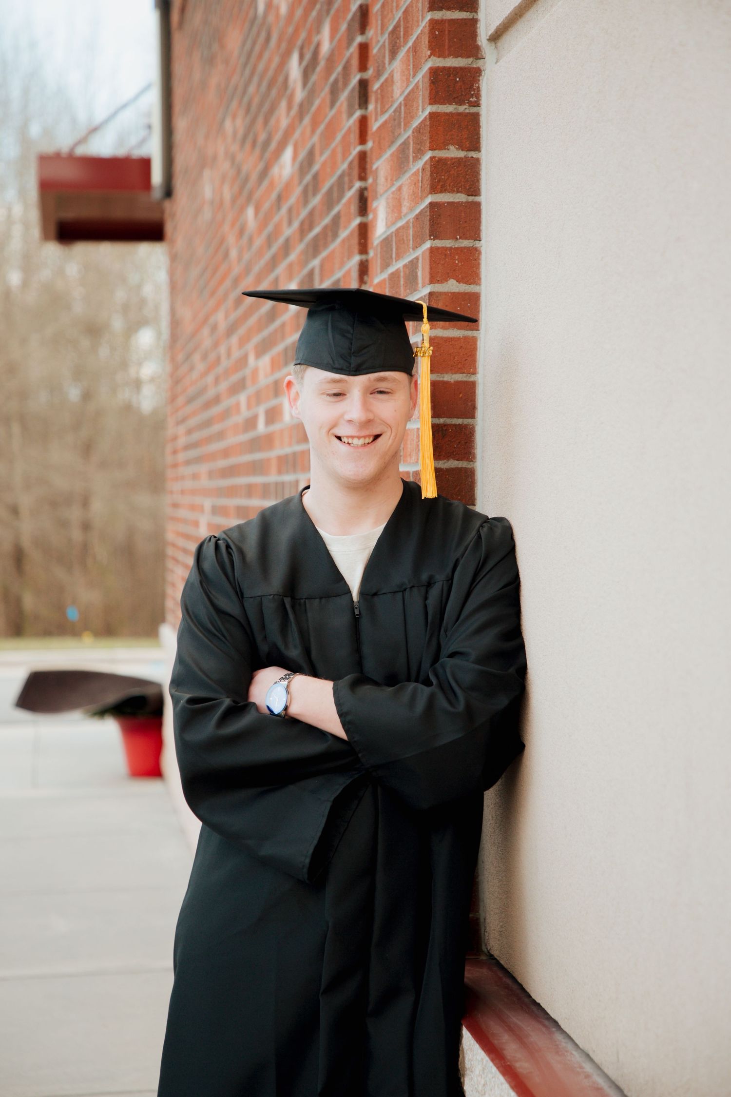 Graduate in black cap and gown smiling with arms crossed against brick wall.