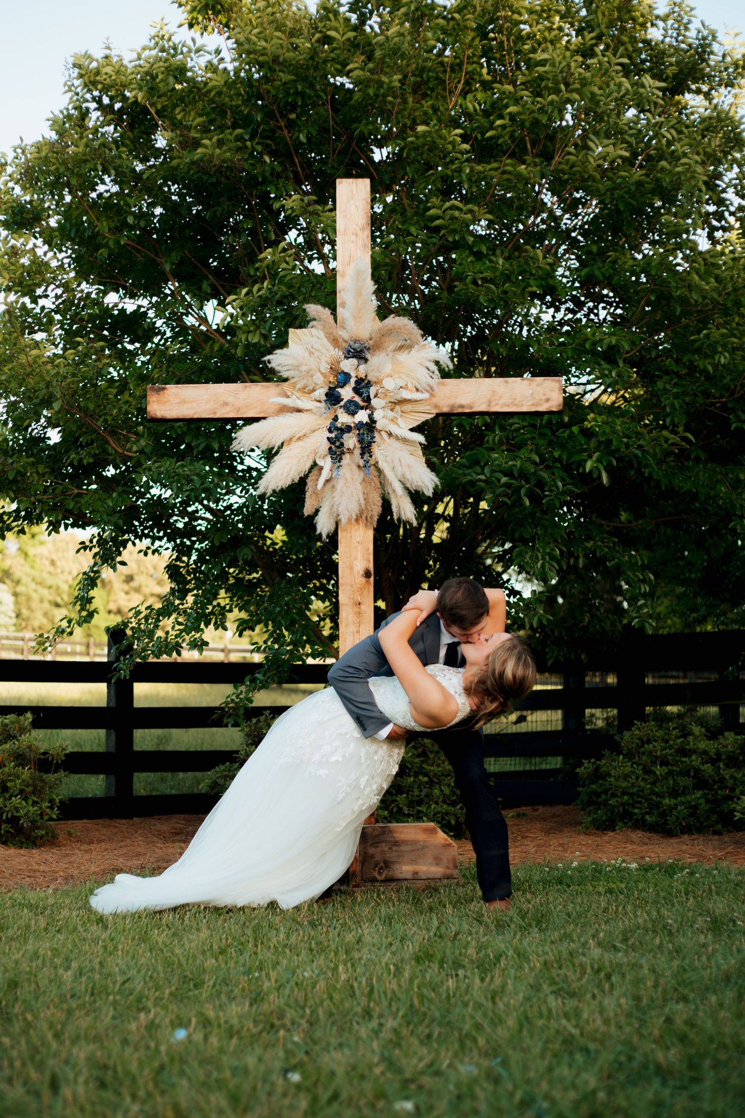 Groom dips bride in a romantic kiss in front of a rustic wooden cross with floral decor at an outdoor farm wedding.