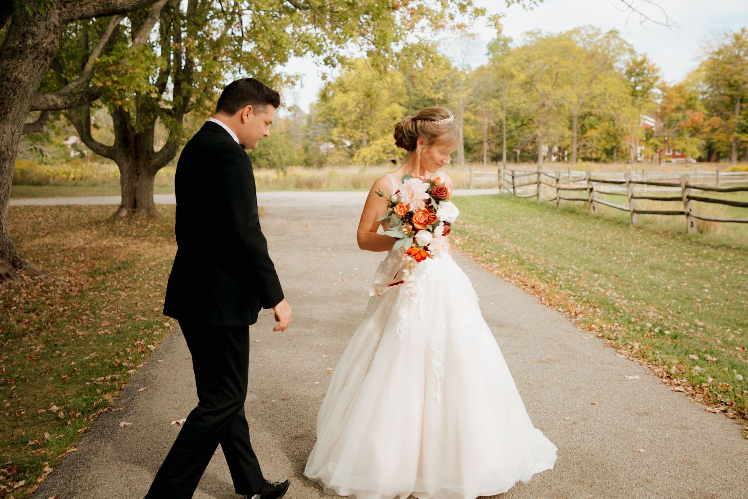 Bride and groom walking together on scenic autumn path, bride holding orange bouquet.