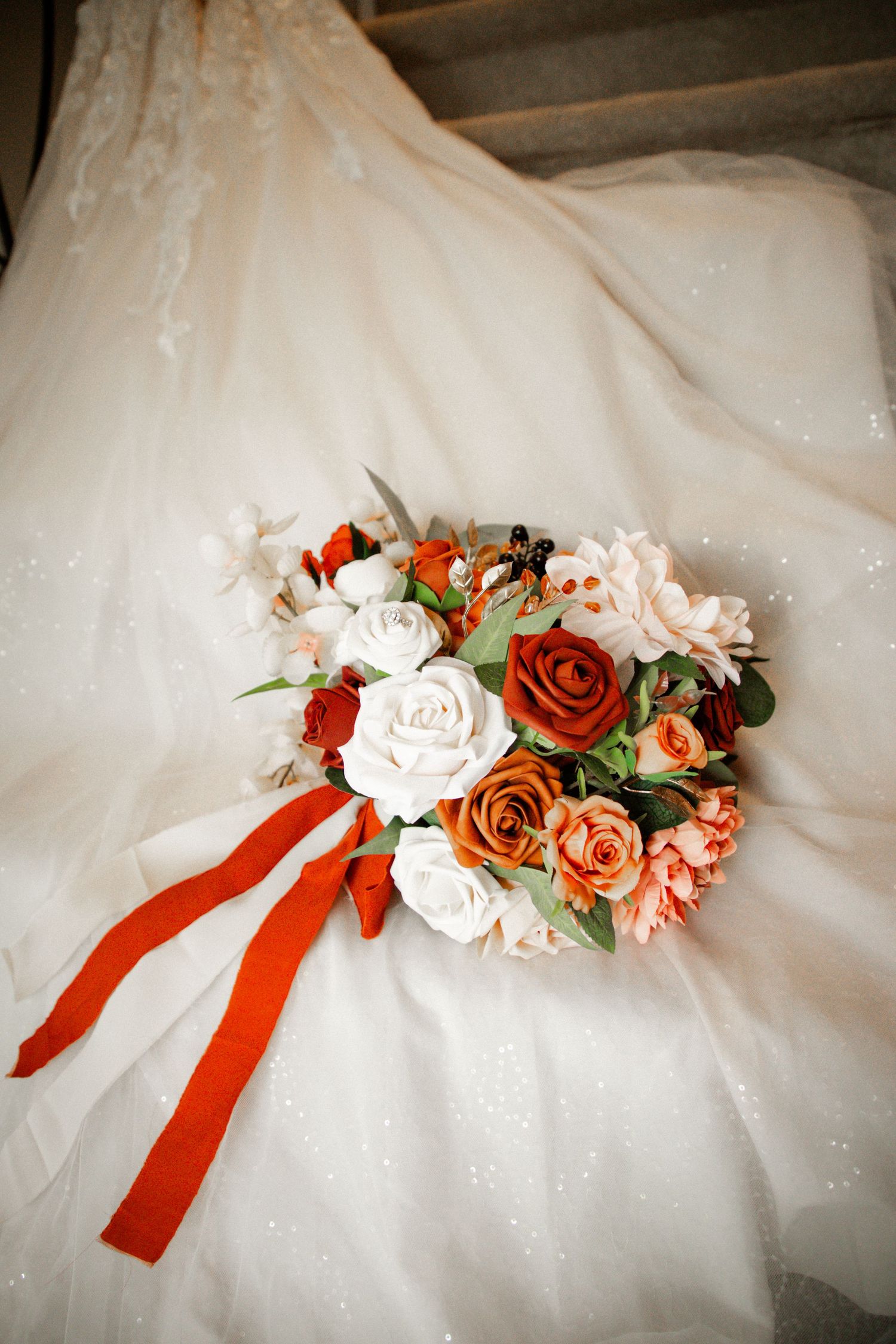 Bridal bouquet with red, orange, and white roses tied with red ribbon on a white lace wedding dress.