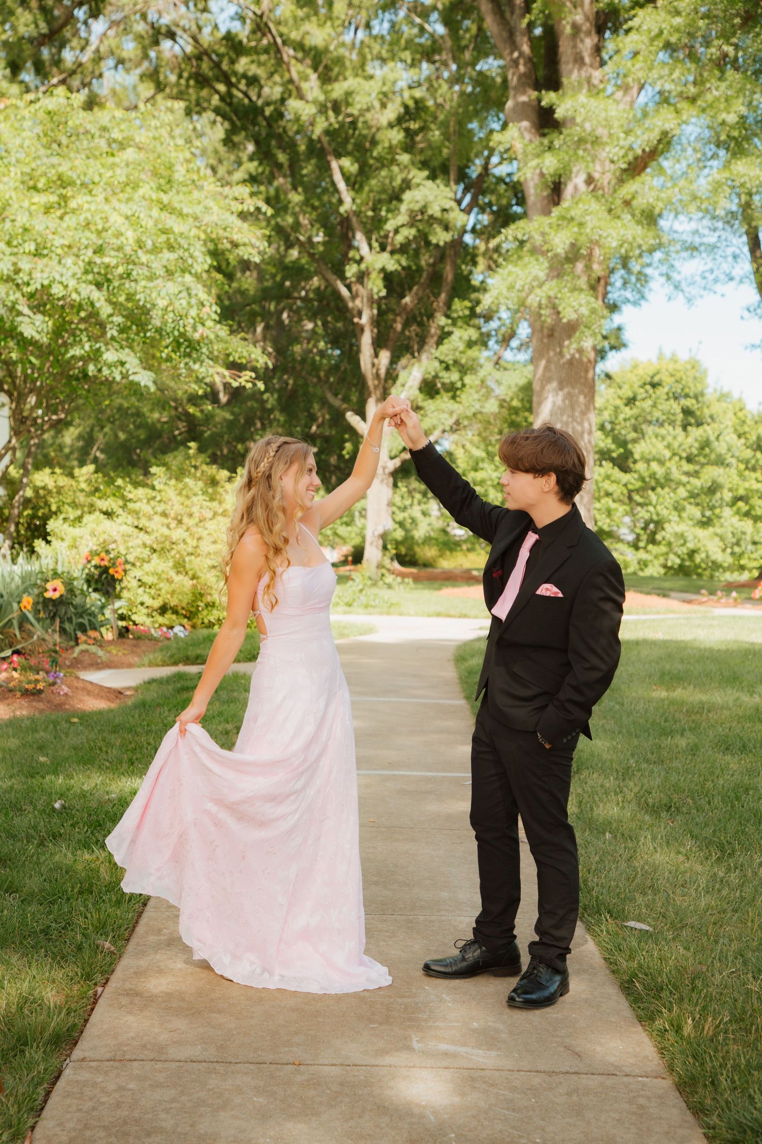 Teen prom couple dancing outdoors, boy in black suit with pink tie spinning girl in pink gown.