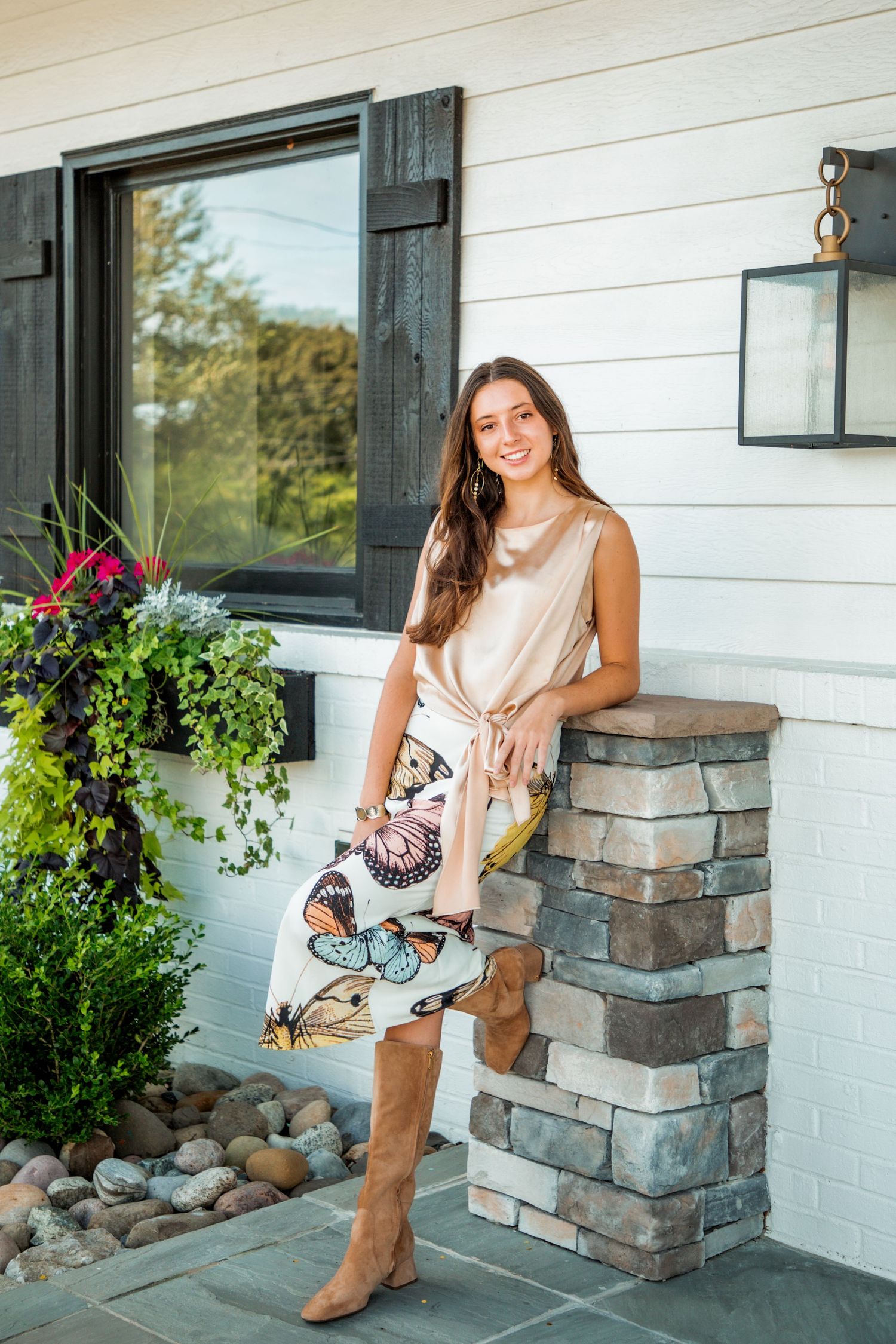 Stylish young woman in gold top and butterfly skirt with tan boots posing on stone porch.