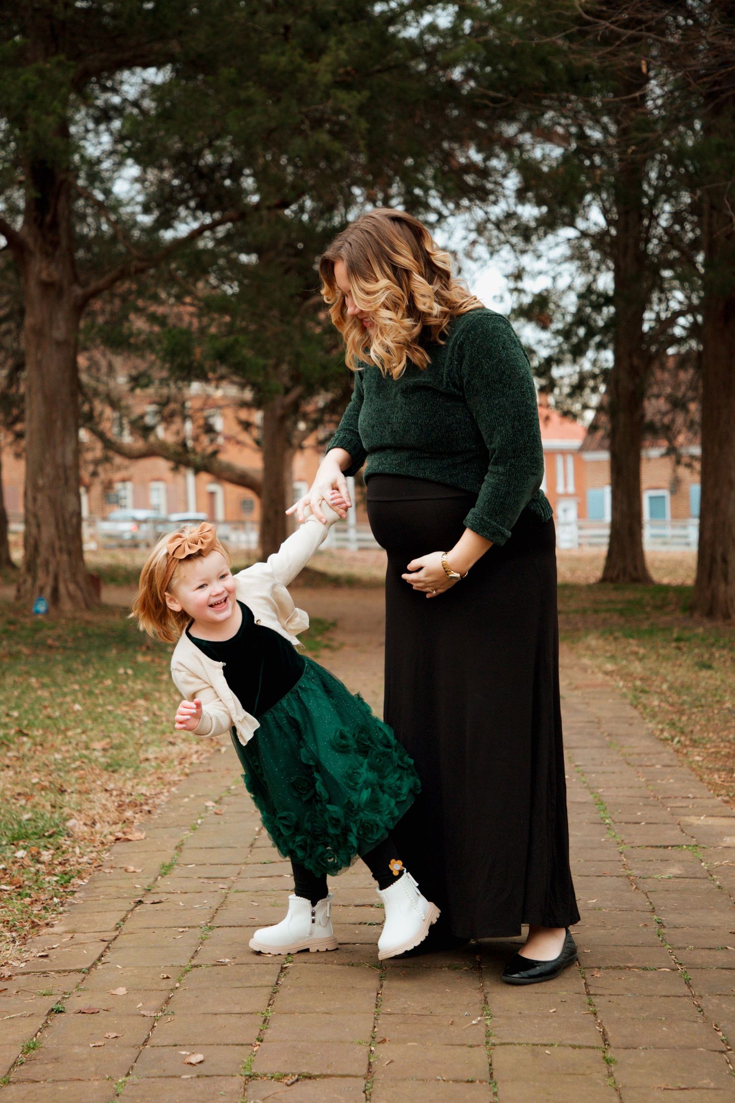 Pregnant mother in green sweater holds hands with joyful red-haired toddler in green dress on brick path.