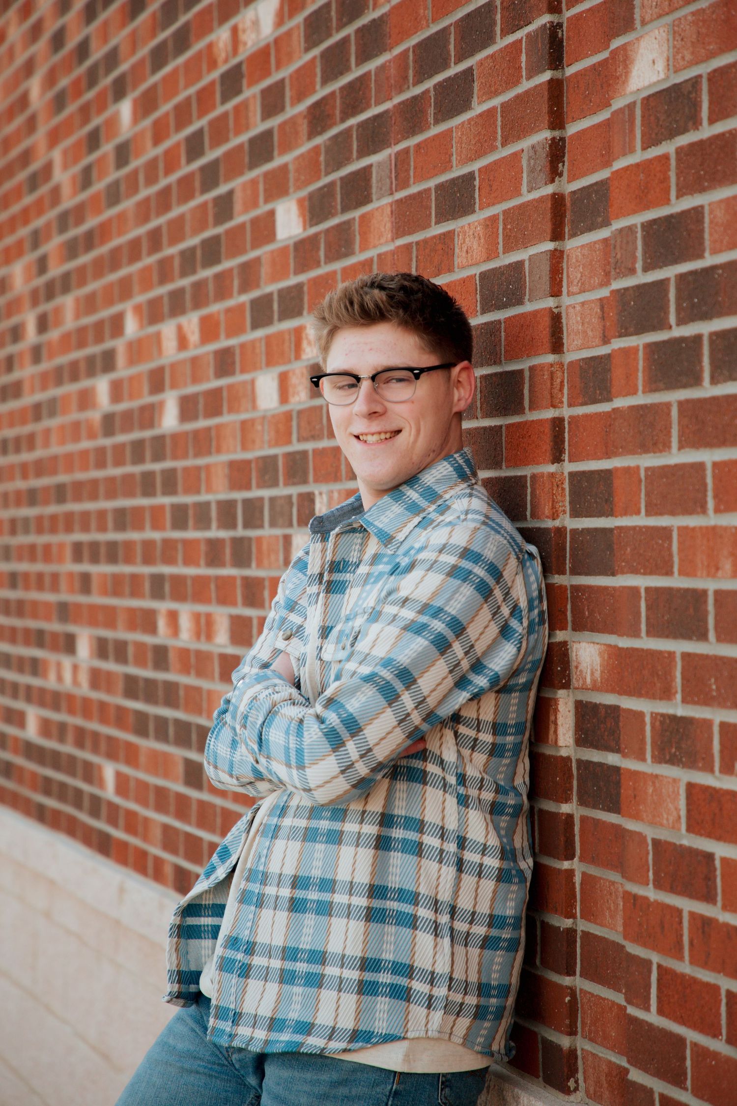 Smiling teen in glasses and blue plaid shirt posing against red brick wall.
