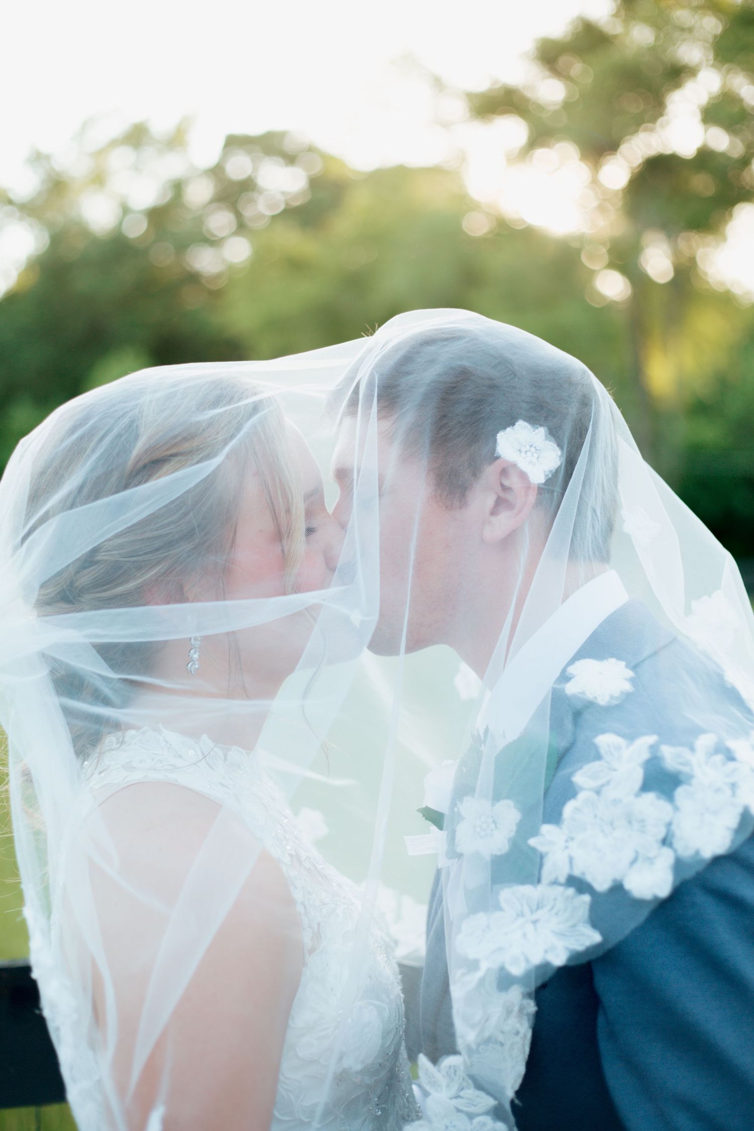 Bride and groom share a kiss hidden beneath flowing white bridal veil outdoors.