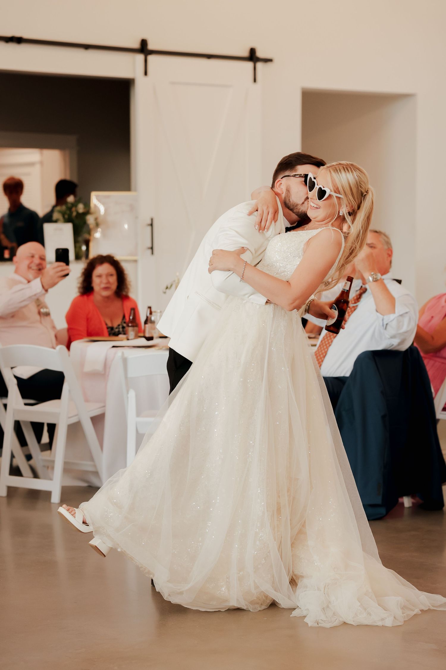 Groom lifting bride during first dance at wedding reception, bride wearing heart-shaped sunglasses, guests watching in background.