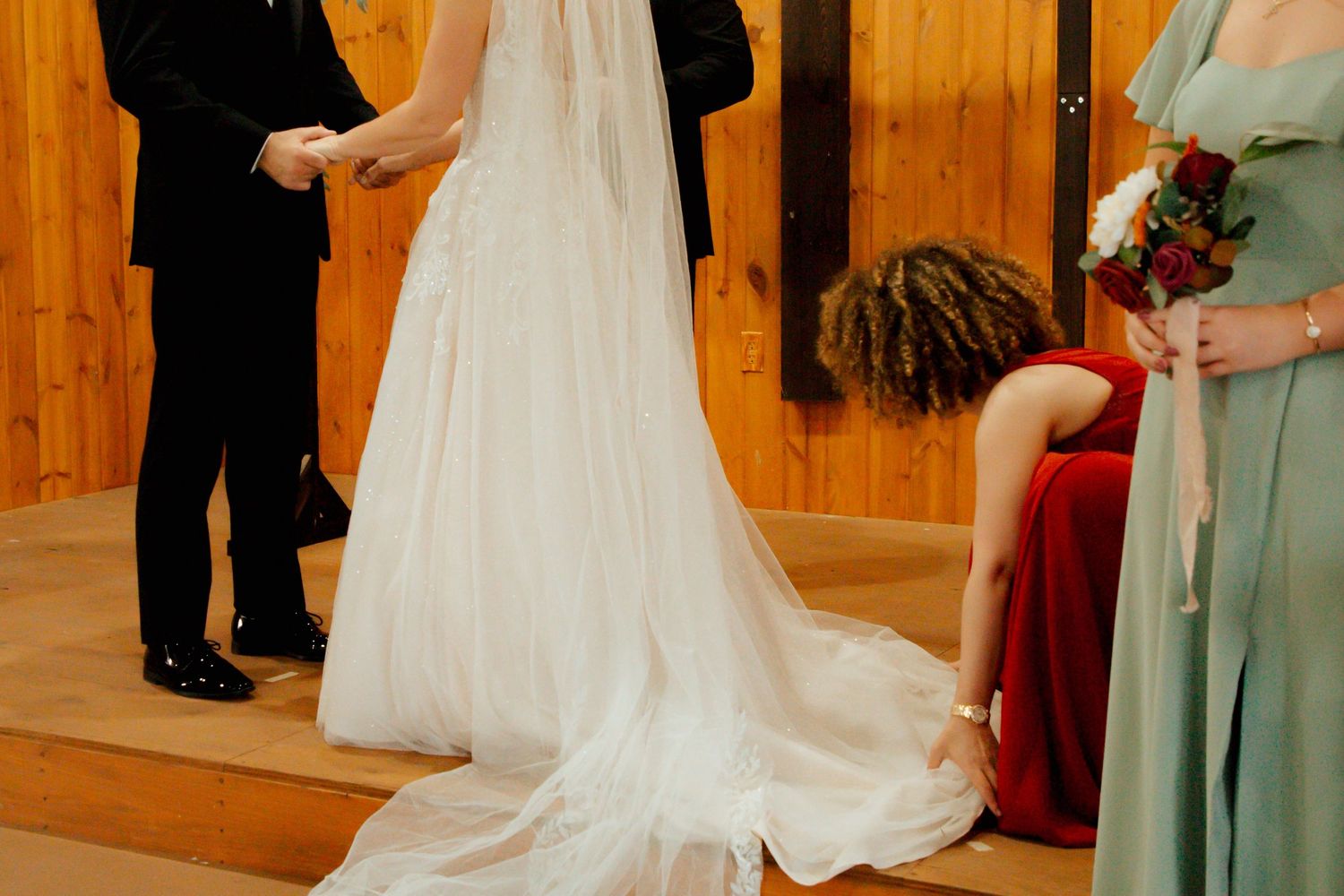 A bridesmaid in a sage green dress adjusts the brides flowing white train during the wedding ceremony.