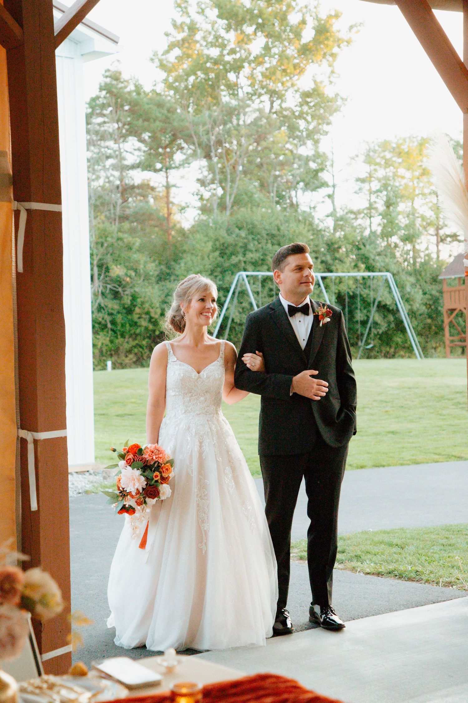 Bride and groom walk arm-in-arm through barn venue entrance with lush greenery behind.