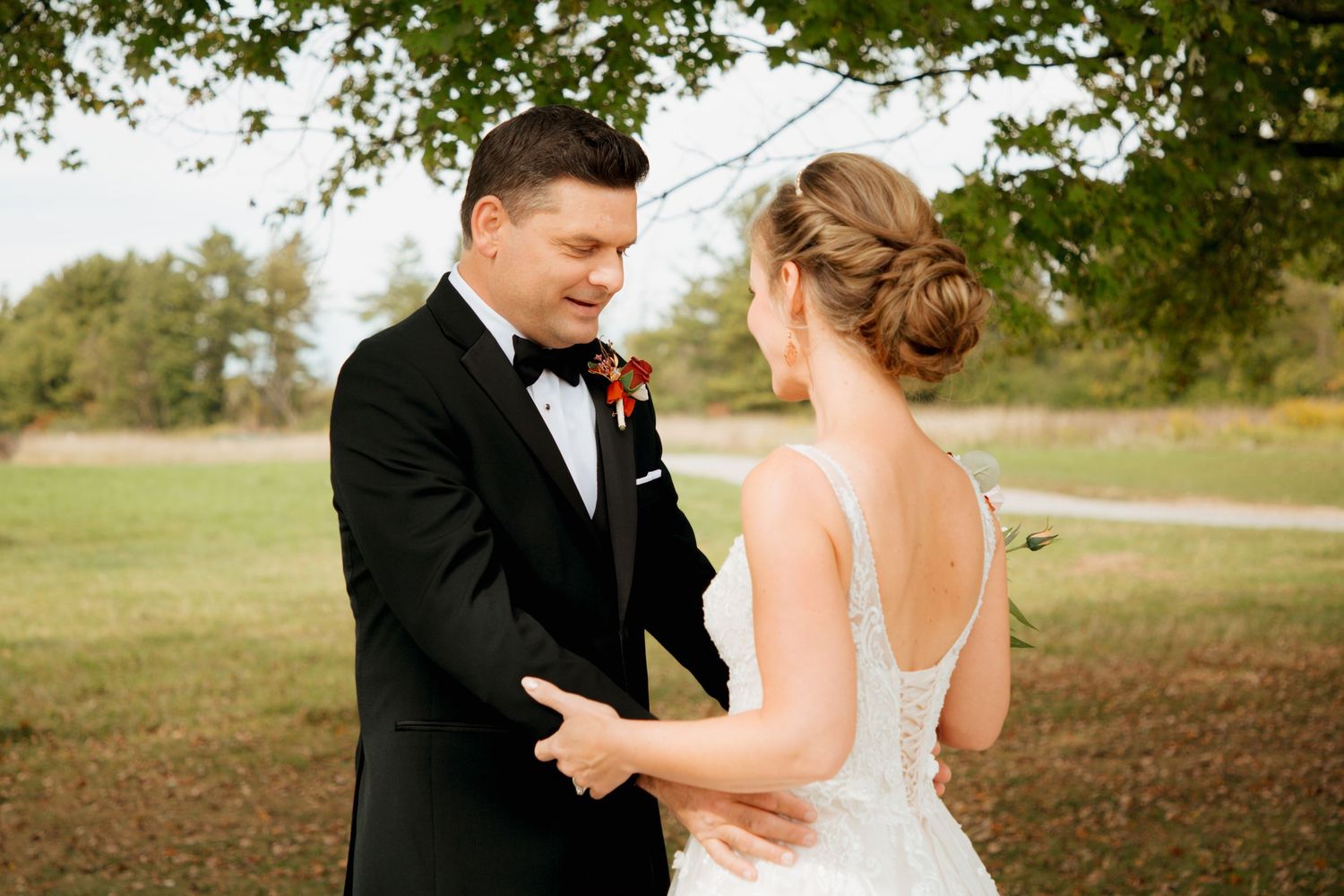 Groom smiles joyfully as he holds brides hands during outdoor first look under a large autumn tree.