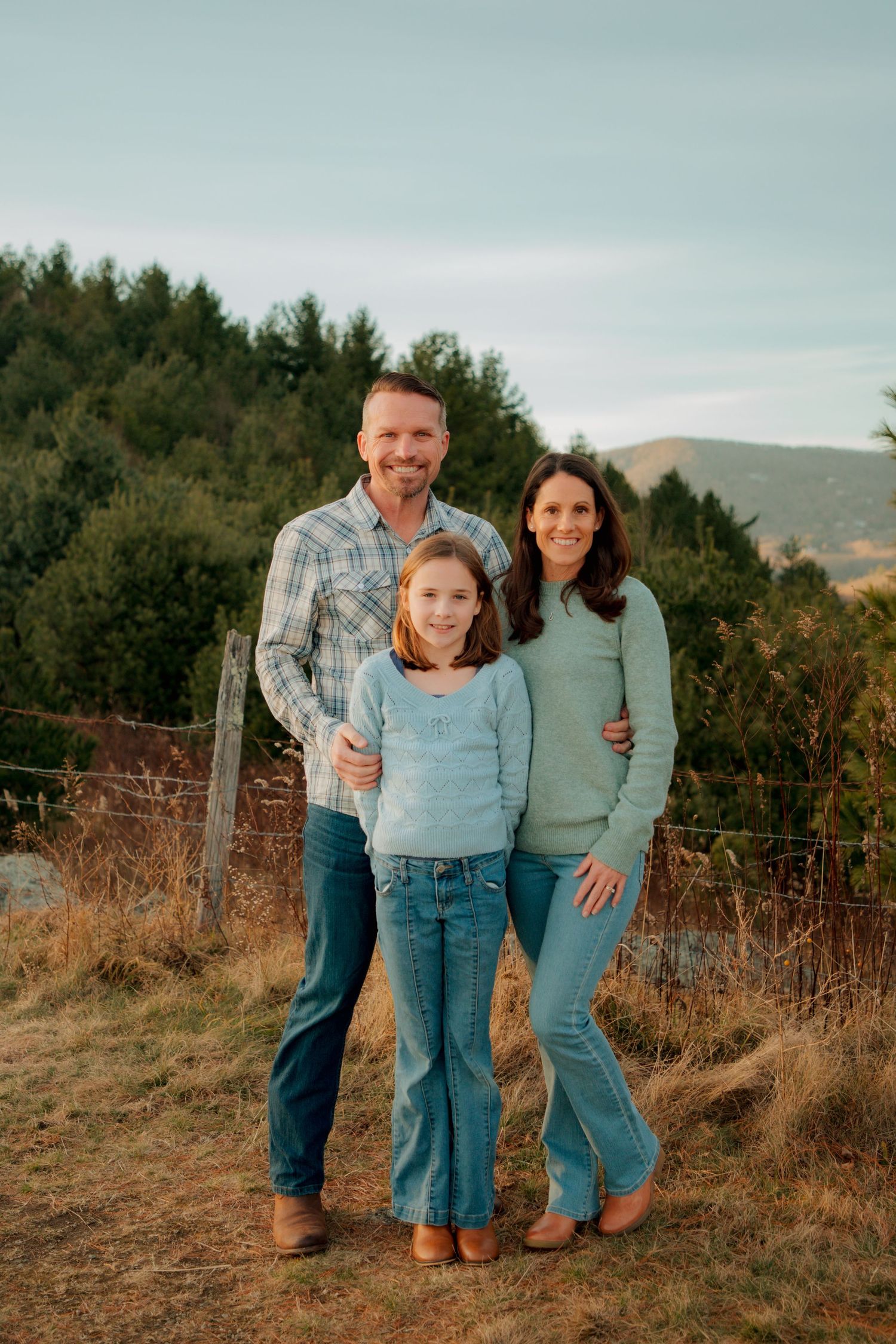 Family of three poses outdoors at golden hour with mountain and forest backdrop in coordinating blue outfits.