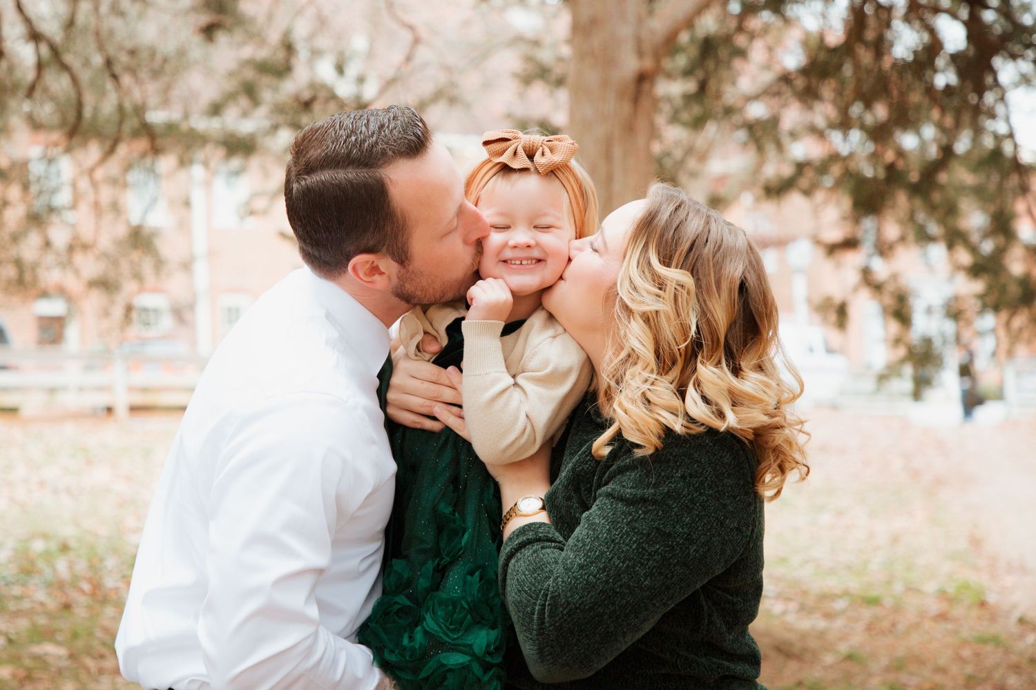 Parents kissing their smiling toddler daughter on cheeks outdoors in autumn park.