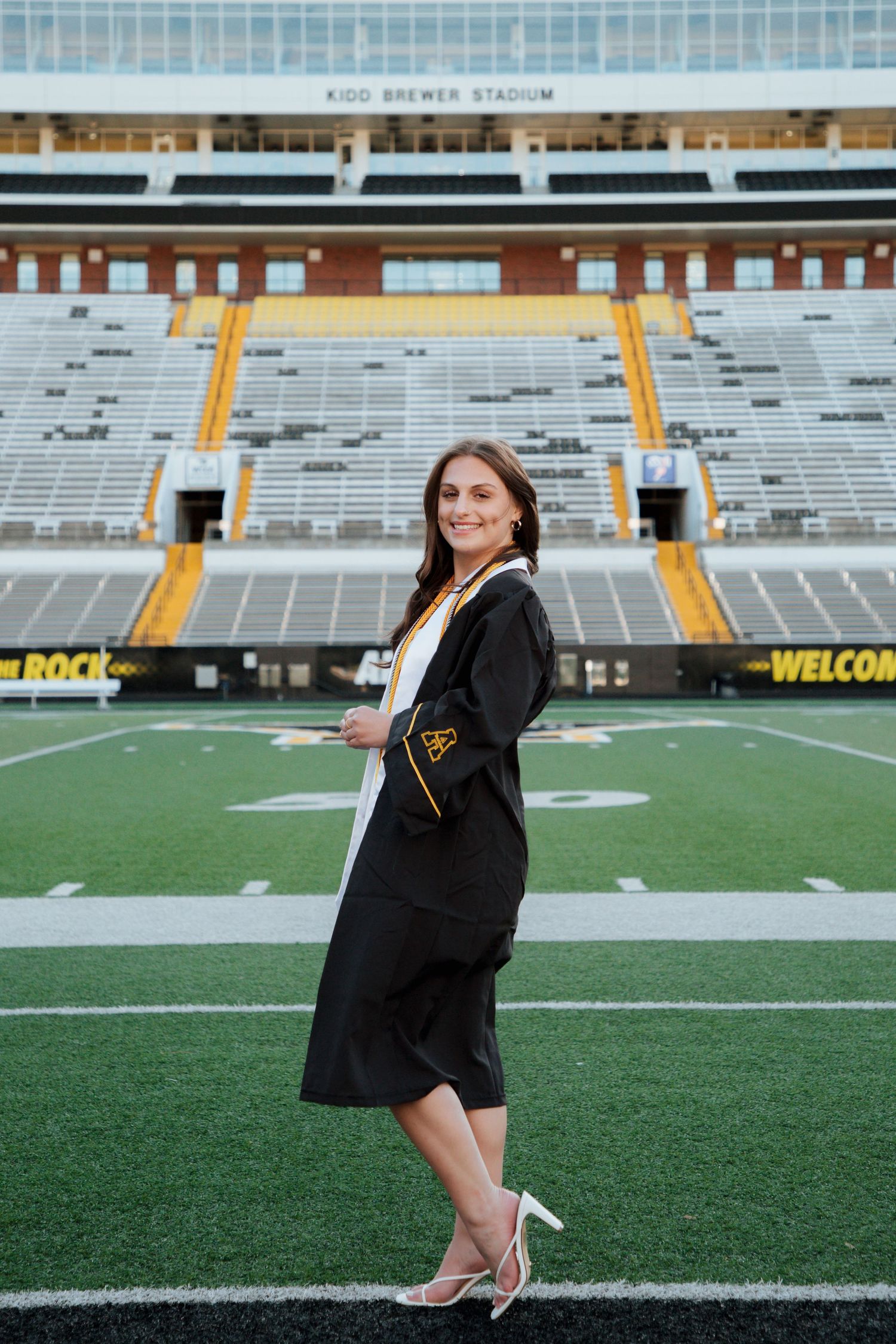 Smiling graduate posing in cap and gown on Kidd Brewer Stadium field at Appalachian State.
