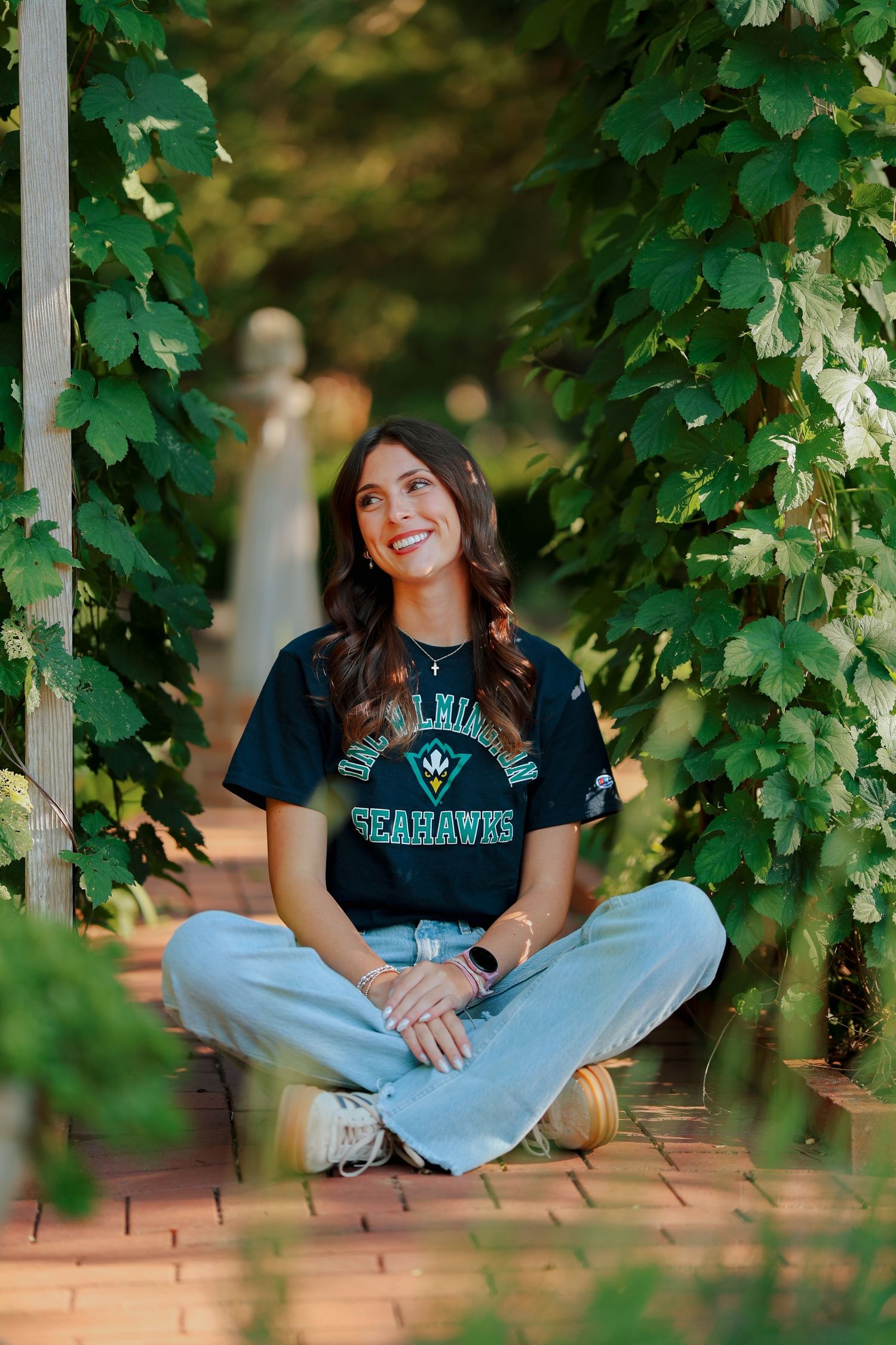 Young woman in UNCW Seahawks shirt sitting cross-legged on brick path beneath a green vine-covered trellis.