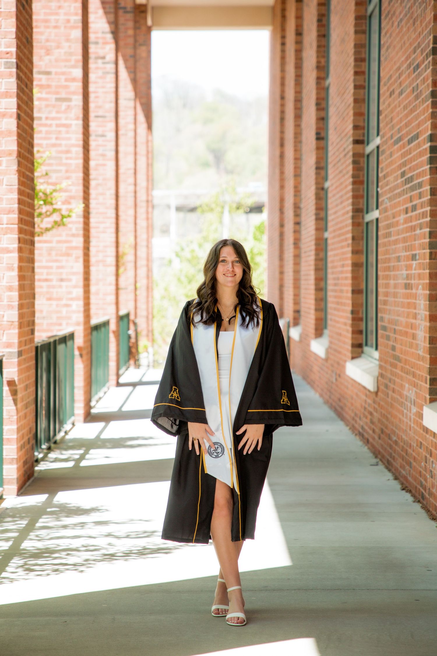 Female graduate in black robe smiling while walking through brick building hallway.