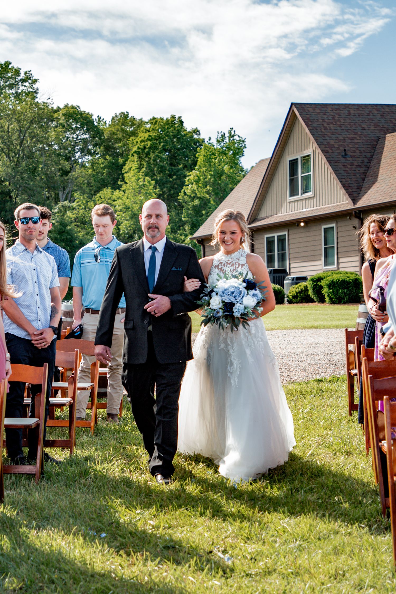 Bride in lace gown with blue bouquet walks down outdoor aisle escorted by father on sunny day.