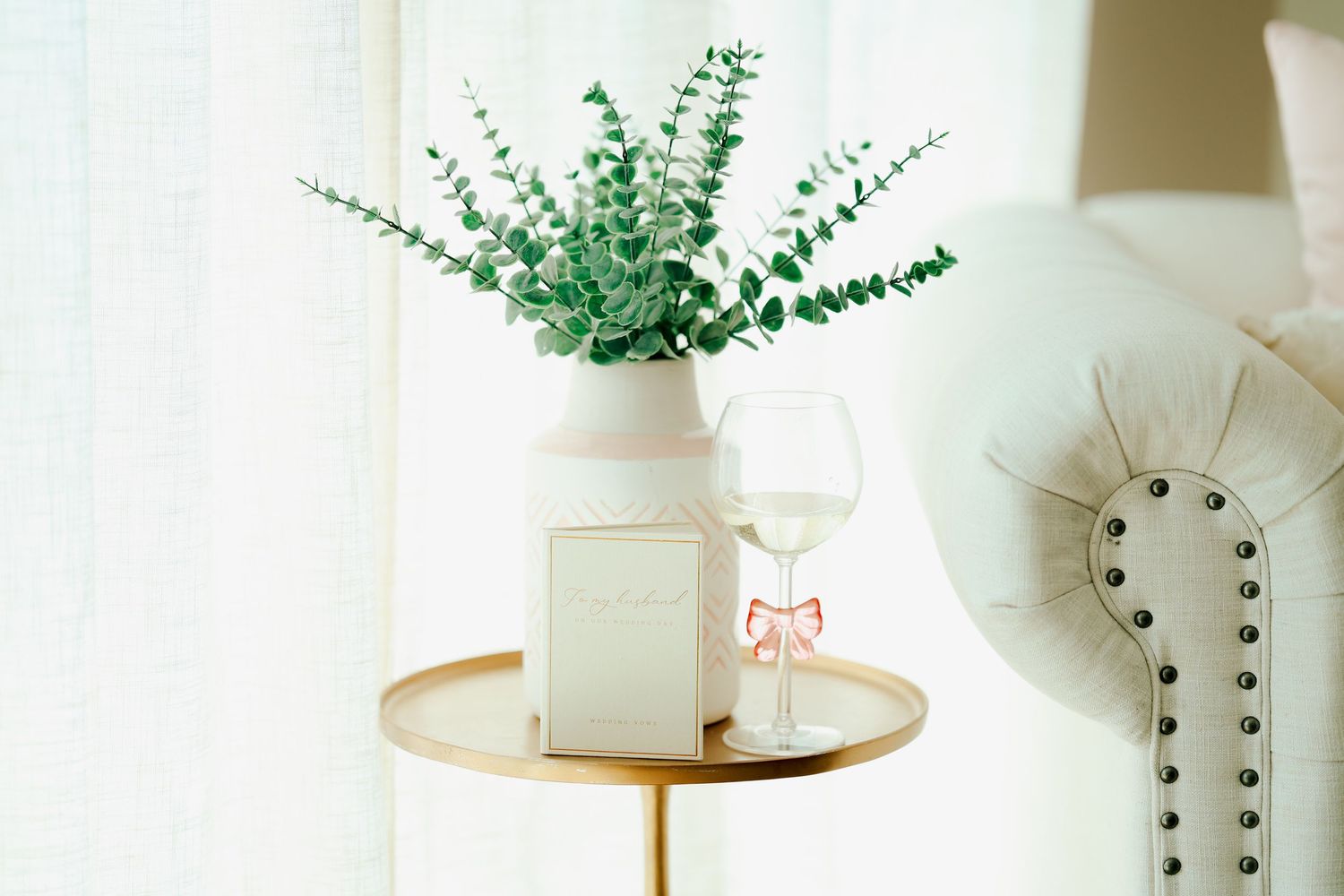 White vase with eucalyptus, wedding program, and wine glass on gold side table beside white chair.