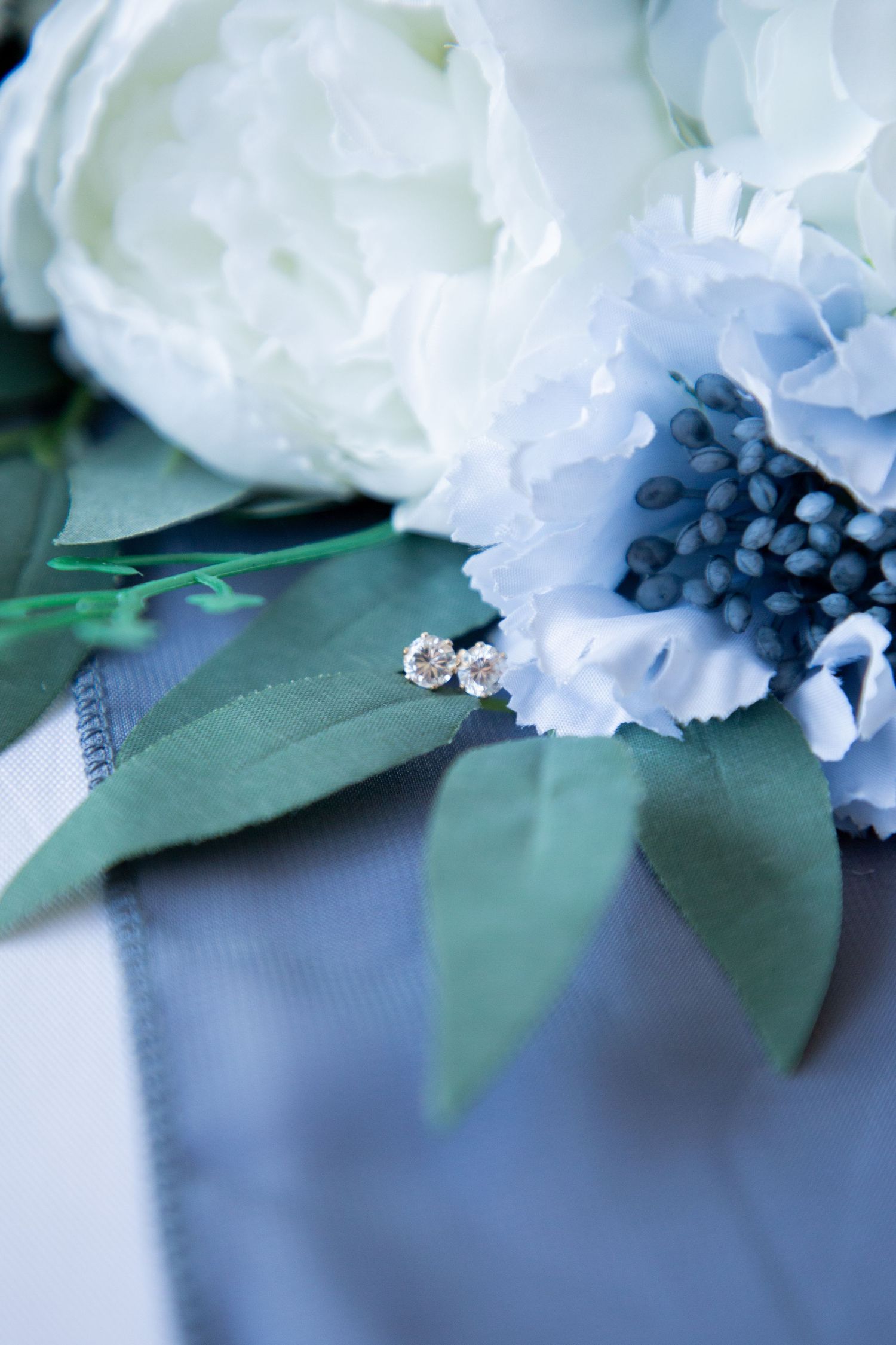 Small diamond stud earrings resting on green leaves beside white and blue flowers on gray fabric.