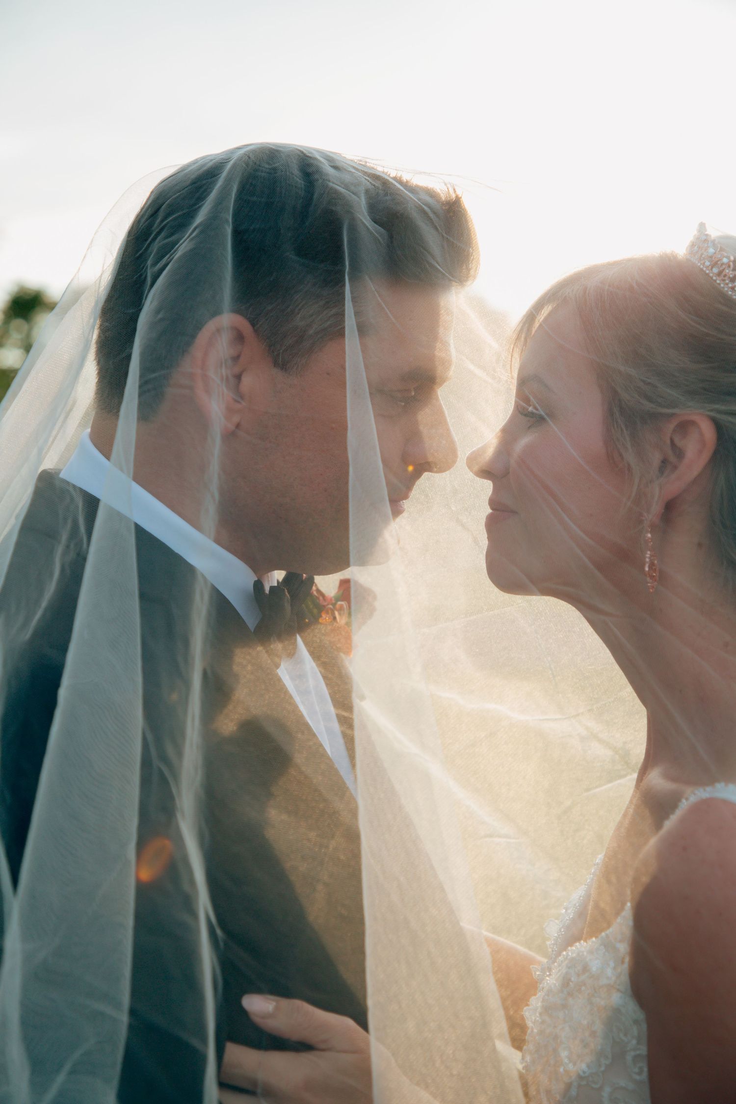Bride and groom lean in for a kiss beneath the flowing veil in a glowing golden hour wedding portrait.