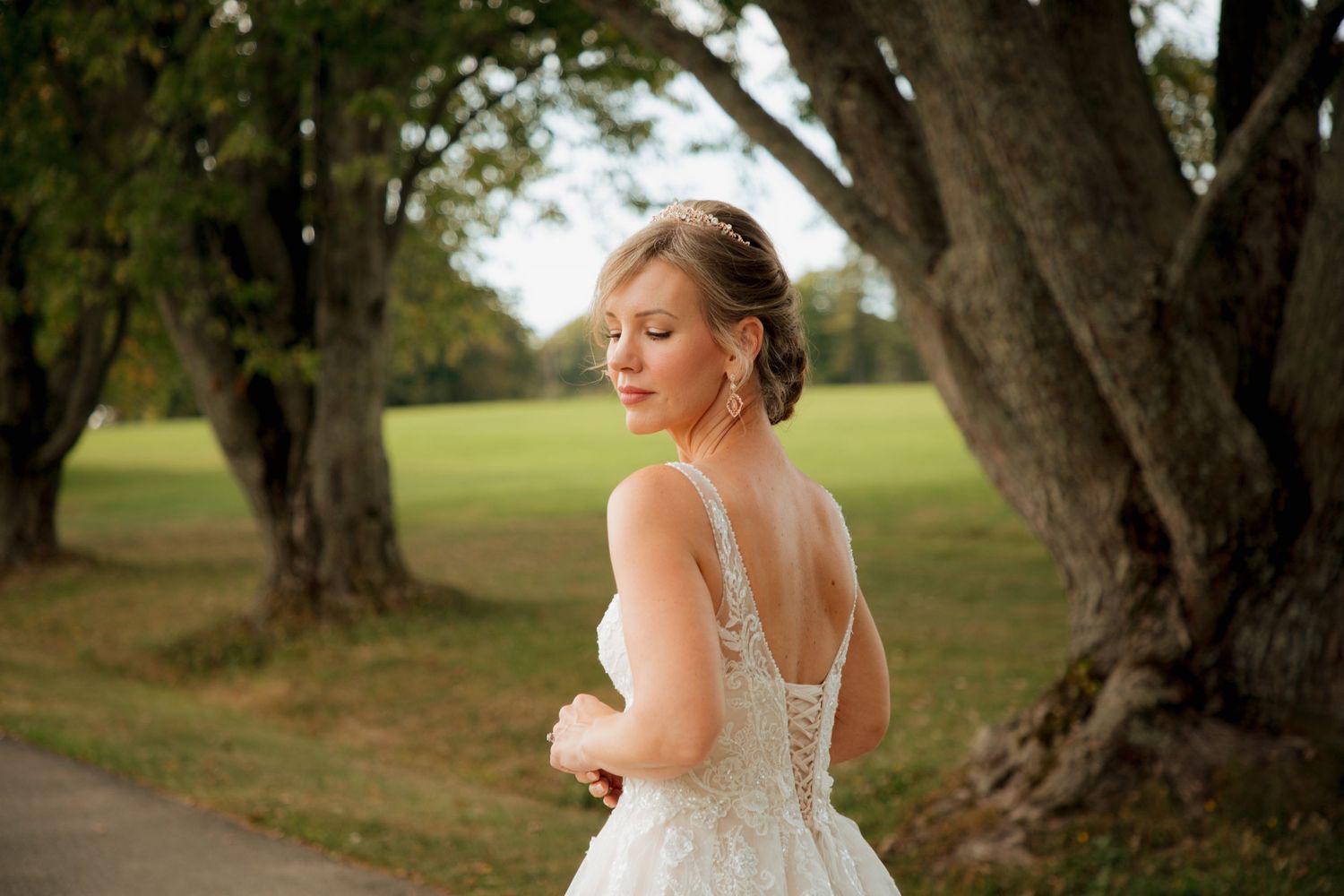 Elegant bride in a lace backless wedding gown poses gracefully among tall trees in a park setting.