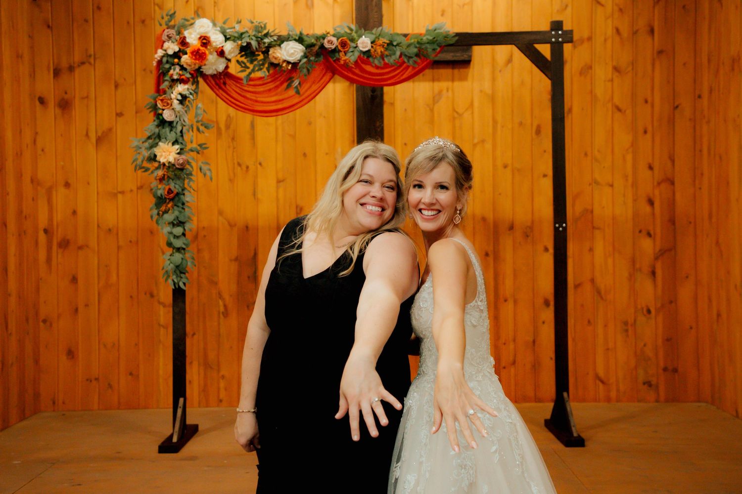 Two women posing happily in front of floral wedding arch, one in bridal gown.