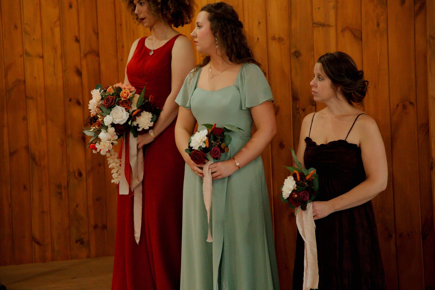 Three bridesmaids in red, sage, and black gowns holding white and burgundy bouquets at ceremony.