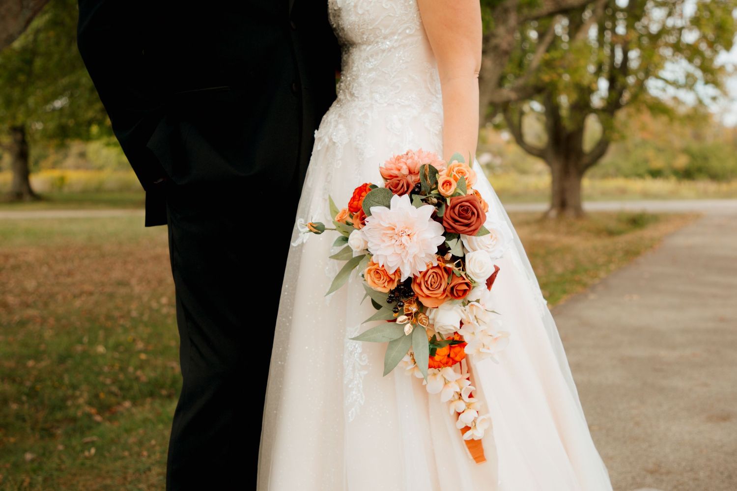Bride holds cascading fall bouquet with orange and pink roses in lace wedding gown.