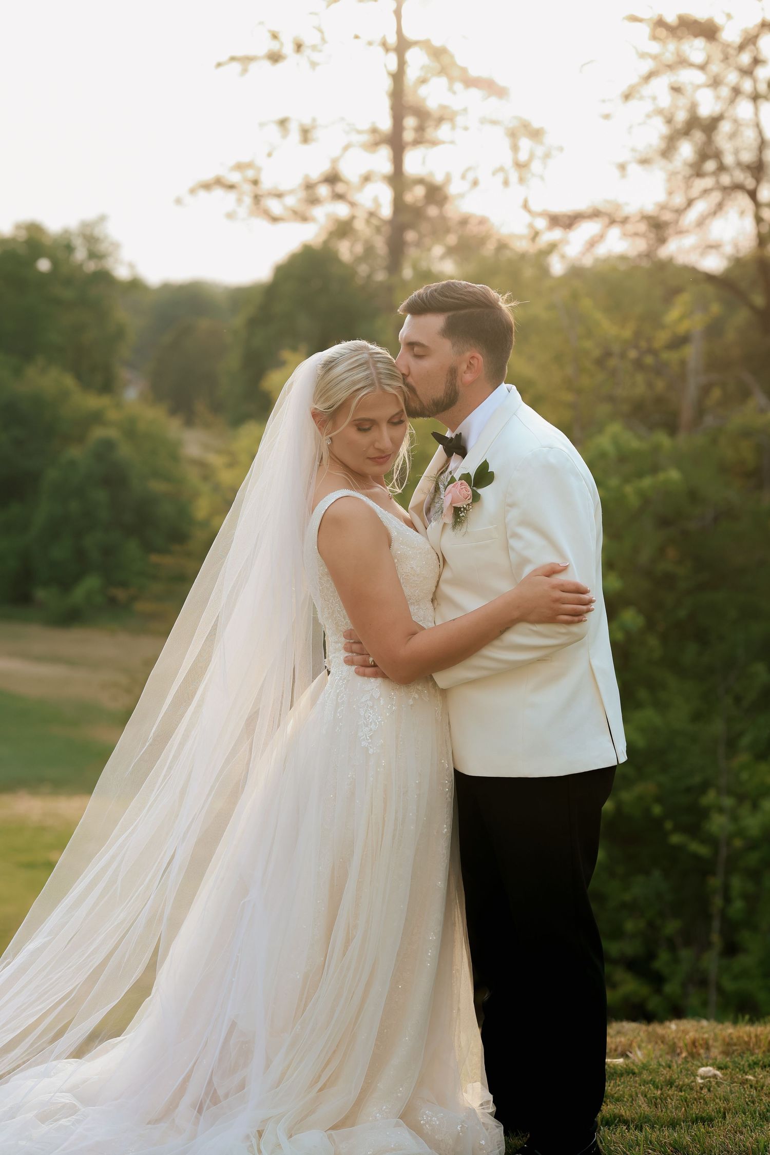 Groom tenderly kisses bride's head during romantic sunset wedding portrait on green hillside.